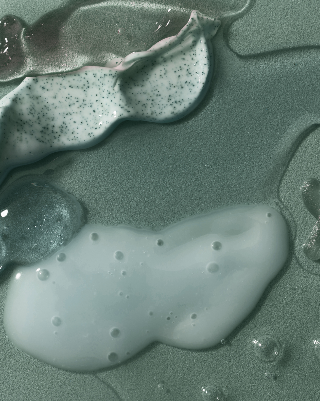 Close-up of two different kinds of skincare creams and serums with bubbles and specks, spread on a textured green surface.