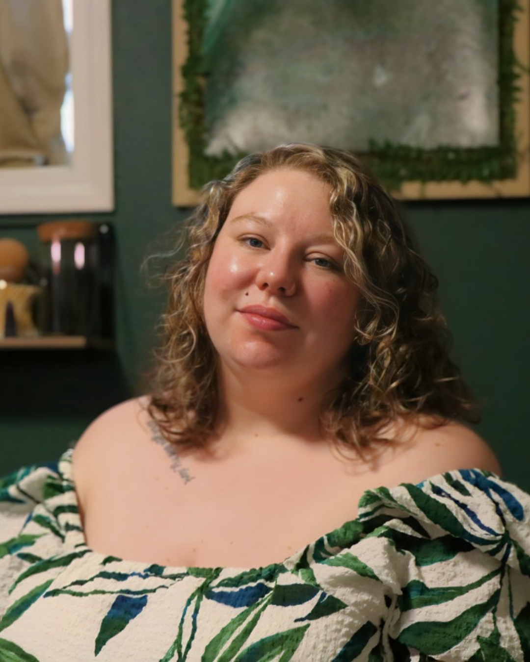 Sarah Gajewski with curly hair and fair skin, wearing a green and white patterned off-shoulder top, sitting indoors with a green wall and framed artwork in the background.