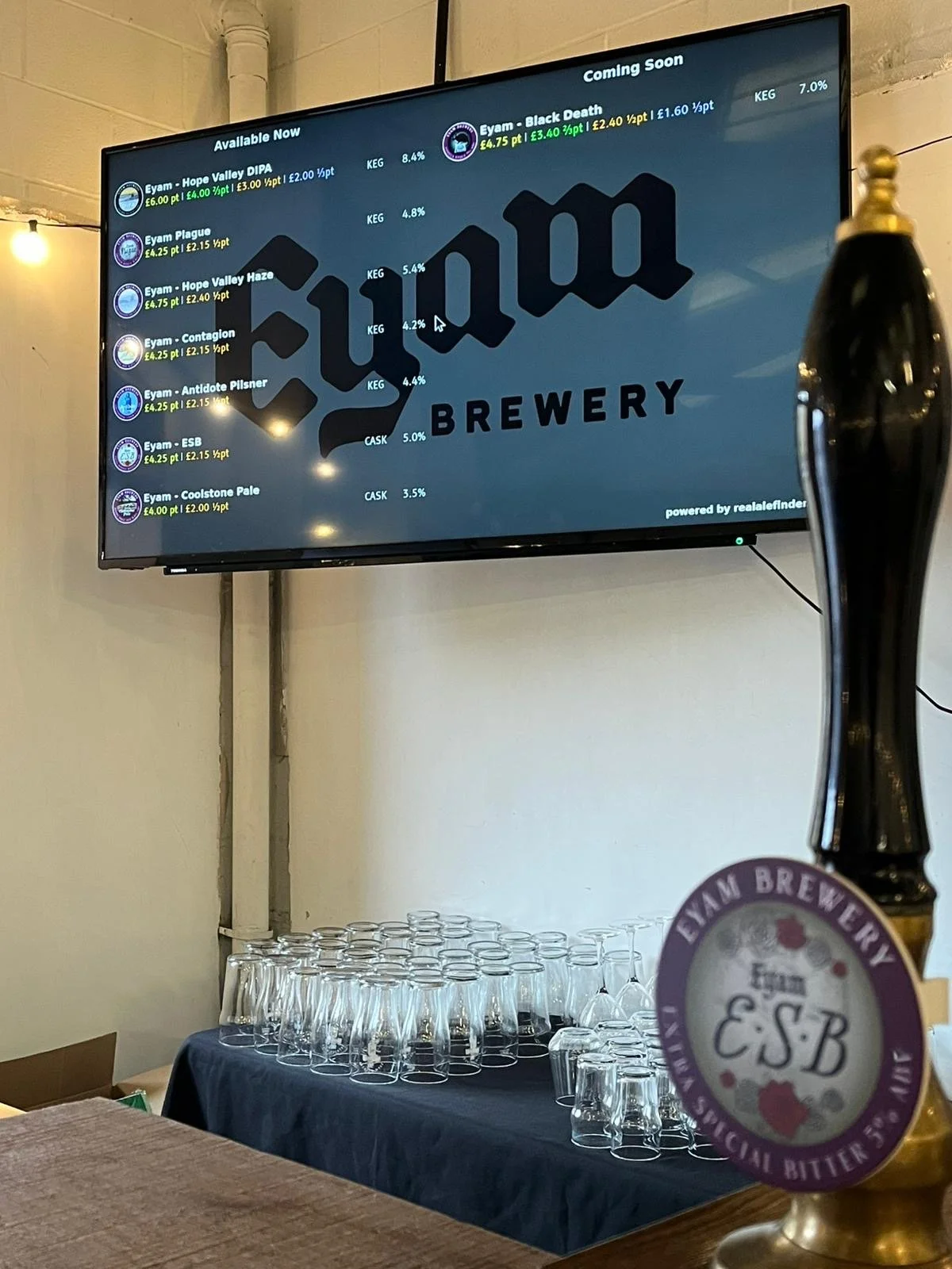 A digital menu board displaying available beer options at a brewery, with glasses arranged on a table below and a beer tap in the foreground.