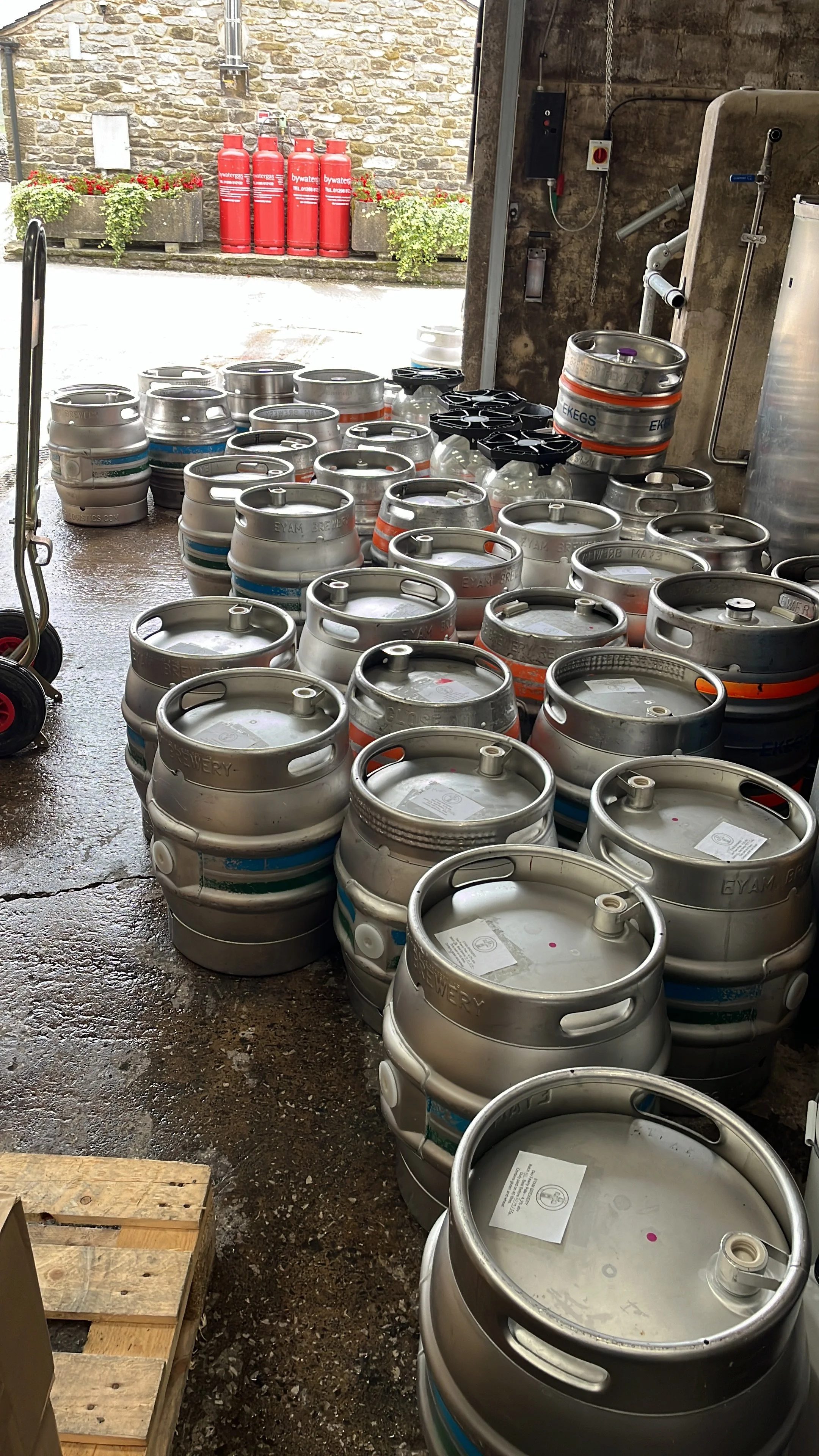 Multiple beer kegs stacked in a cellar, with some on the floor and others on a rack, and fire extinguishers outside the cellar door.
