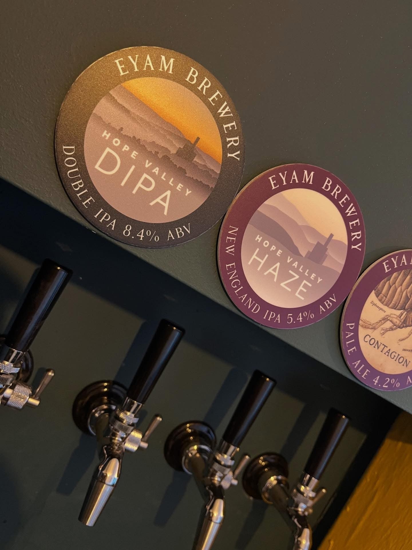 Close-up of beer taps mounted on a bar with circular beer labels advertising Eyam Brewery beers like Hope Valley IPA, Haze Pale Ale, with mountain and sunset backgrounds.
