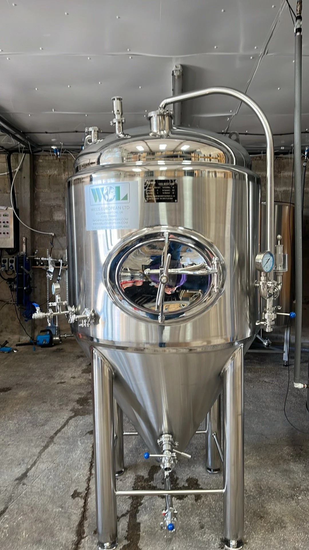 A large stainless steel industrial vessel with a circular port window, gauges, valves, and pipes, situated in a workshop with concrete and brick walls.