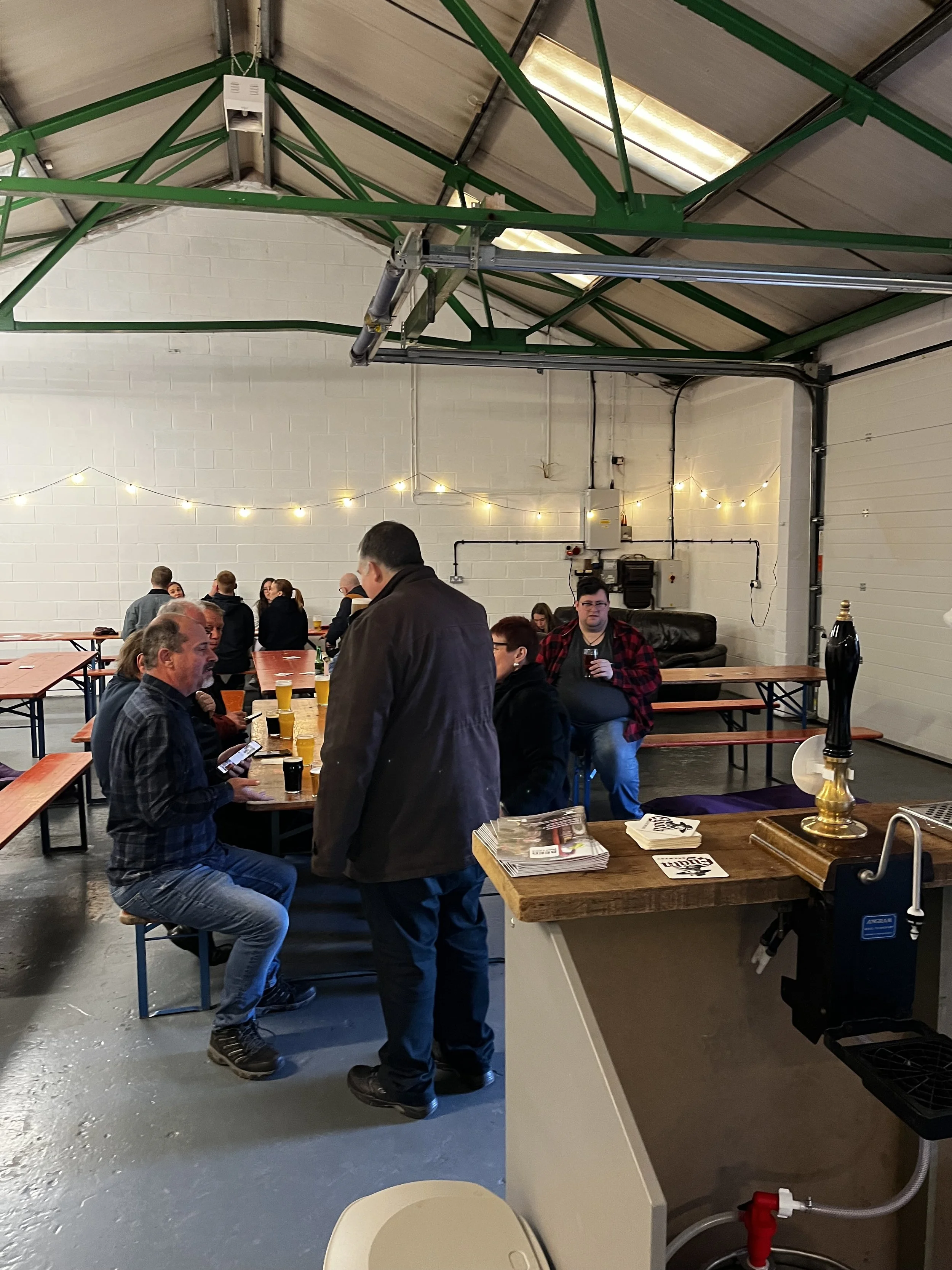 Indoor gathering of people at a casual event in a large industrial style room with white brick walls and string lights. Several individuals are seated at tables with drinks, while some people are standing and conversing.