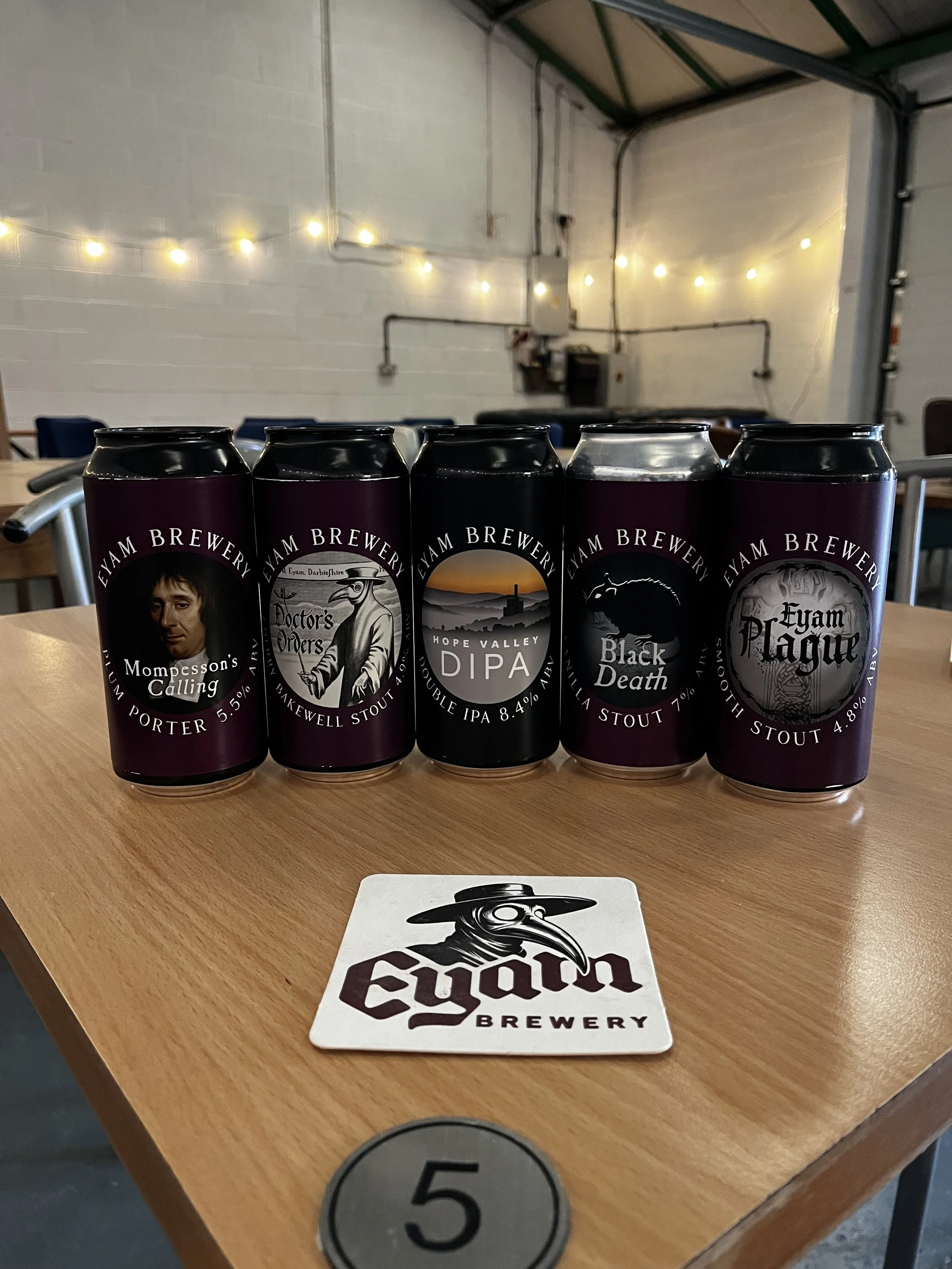 Five cans of Eyam Brewery beer placed on a wooden table in a brewery setting. The cans feature artistic labels with various illustrations. There is a brewery sticker on the table with the Eyam Brewery logo, and a circular sign with the number 5.