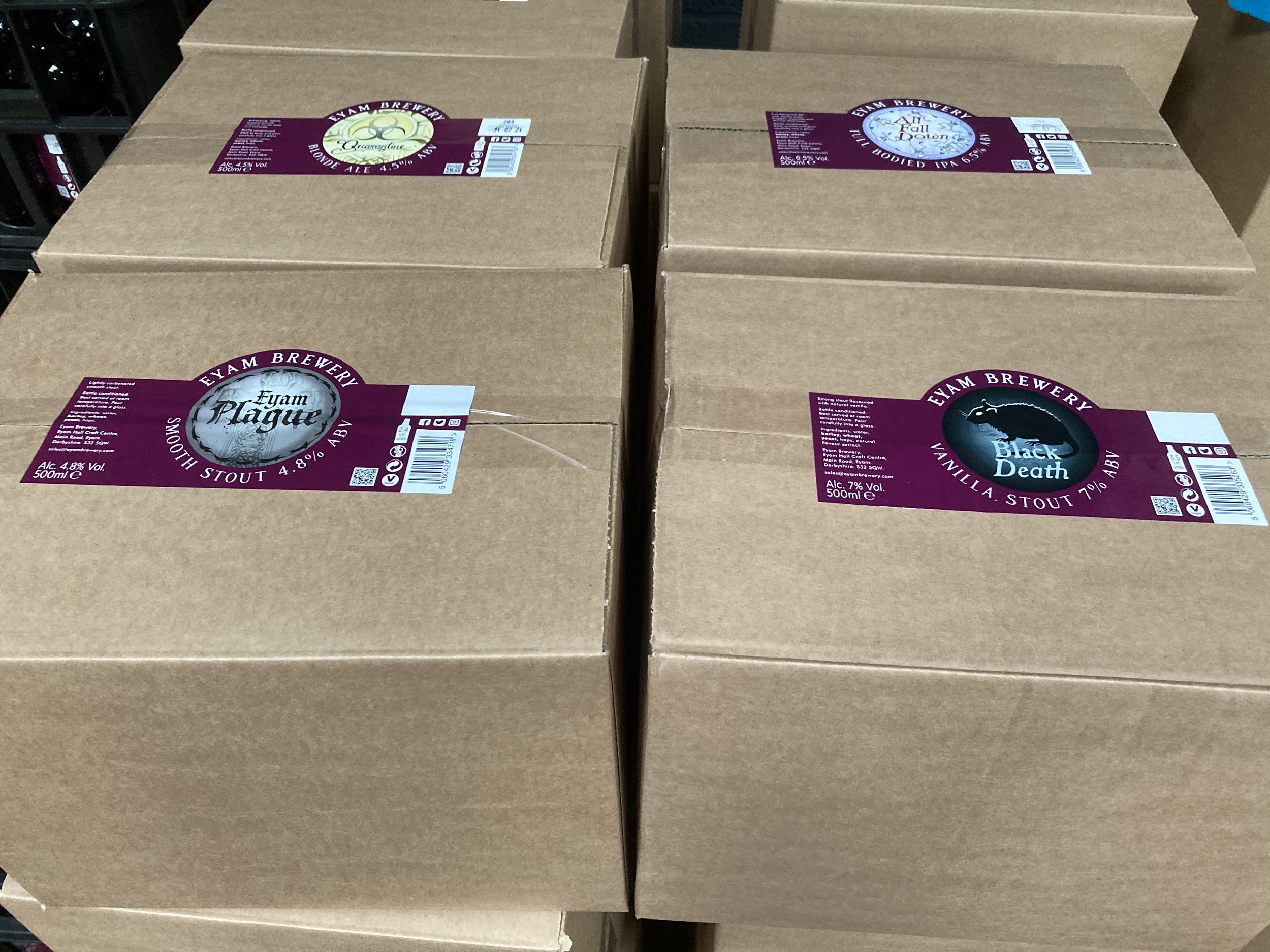 Several cardboard boxes stacked on a shelf, each with a colorful label indicating different types of beer from Eyam Brewery, including Quarantaine Blonde Ale, All Fall Down IPA, Topless Stout, and Black Death Imperial Stout.