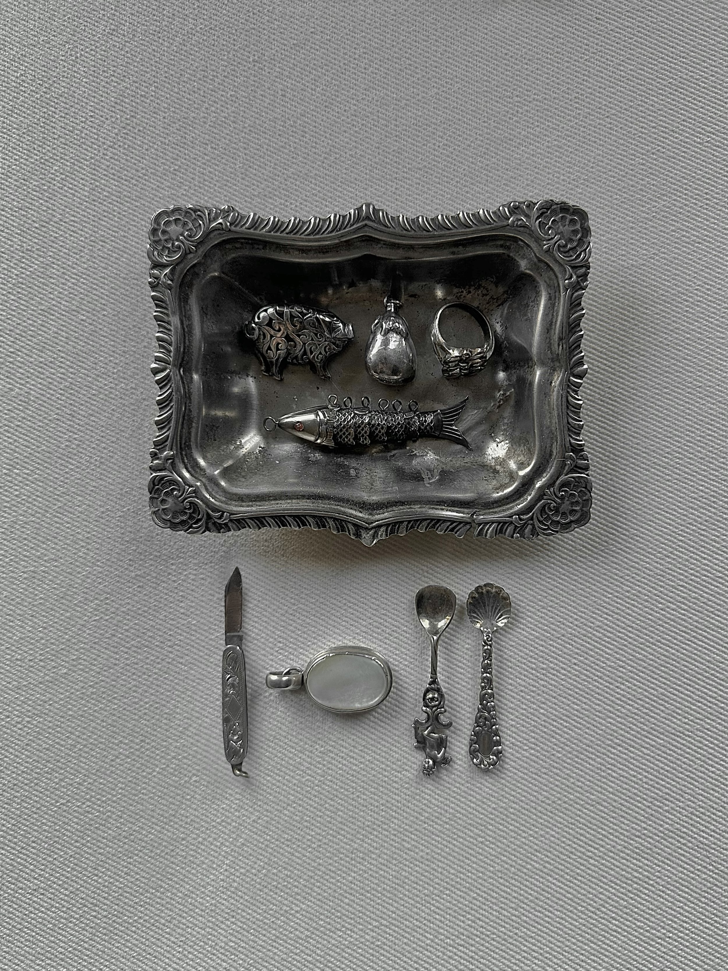 A collection of vintage silver jewelry and tableware on a white textured surface, including rings, a brooch, a spoon, a small knife, a cup, and a fish-shaped pendant, displayed on a decorative silver tray.