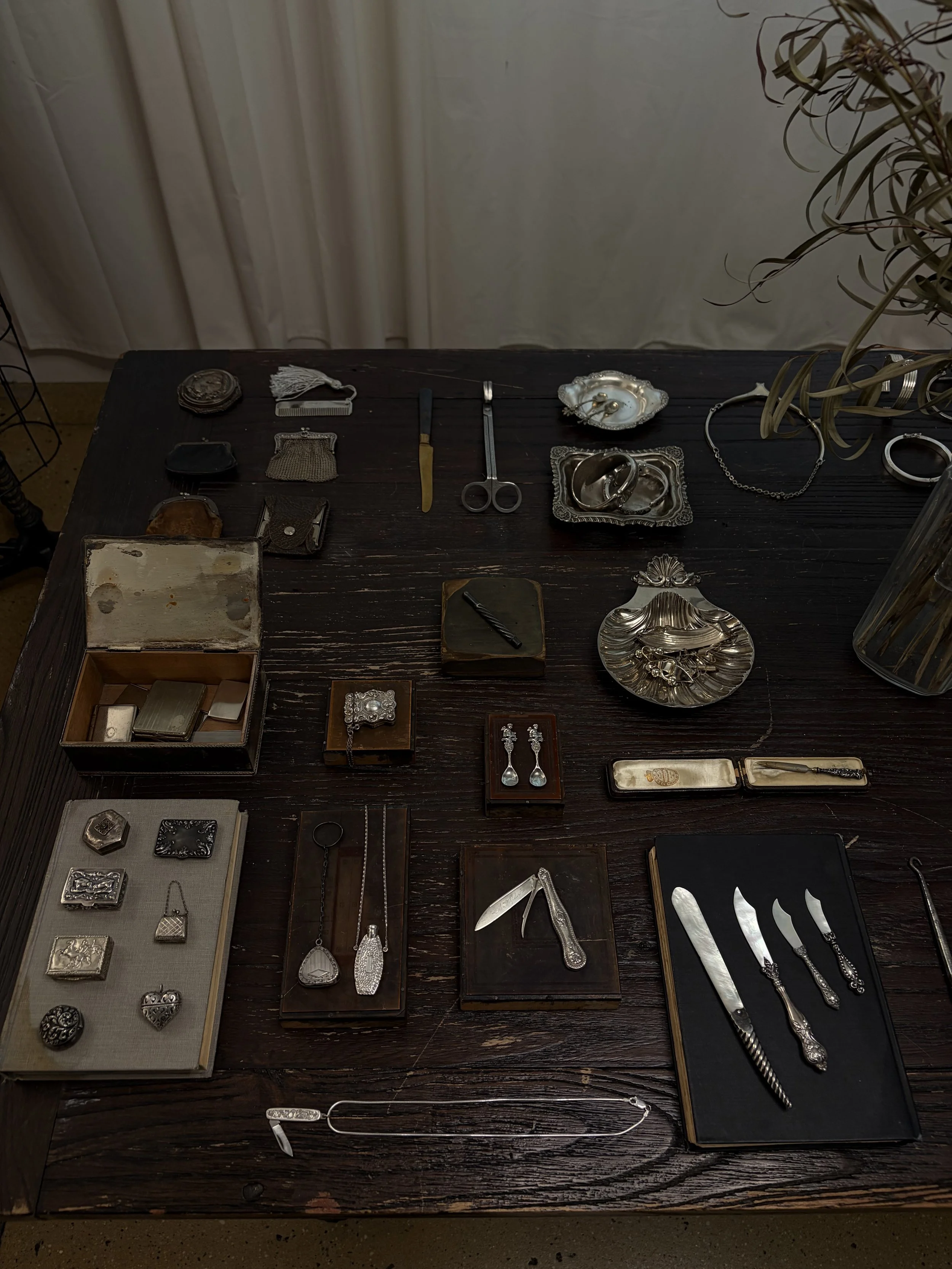 A dark wooden table displaying various pieces of jewelry and silverware, including brooches, rings, earrings, necklaces, and knives, arranged in boxes and trays.