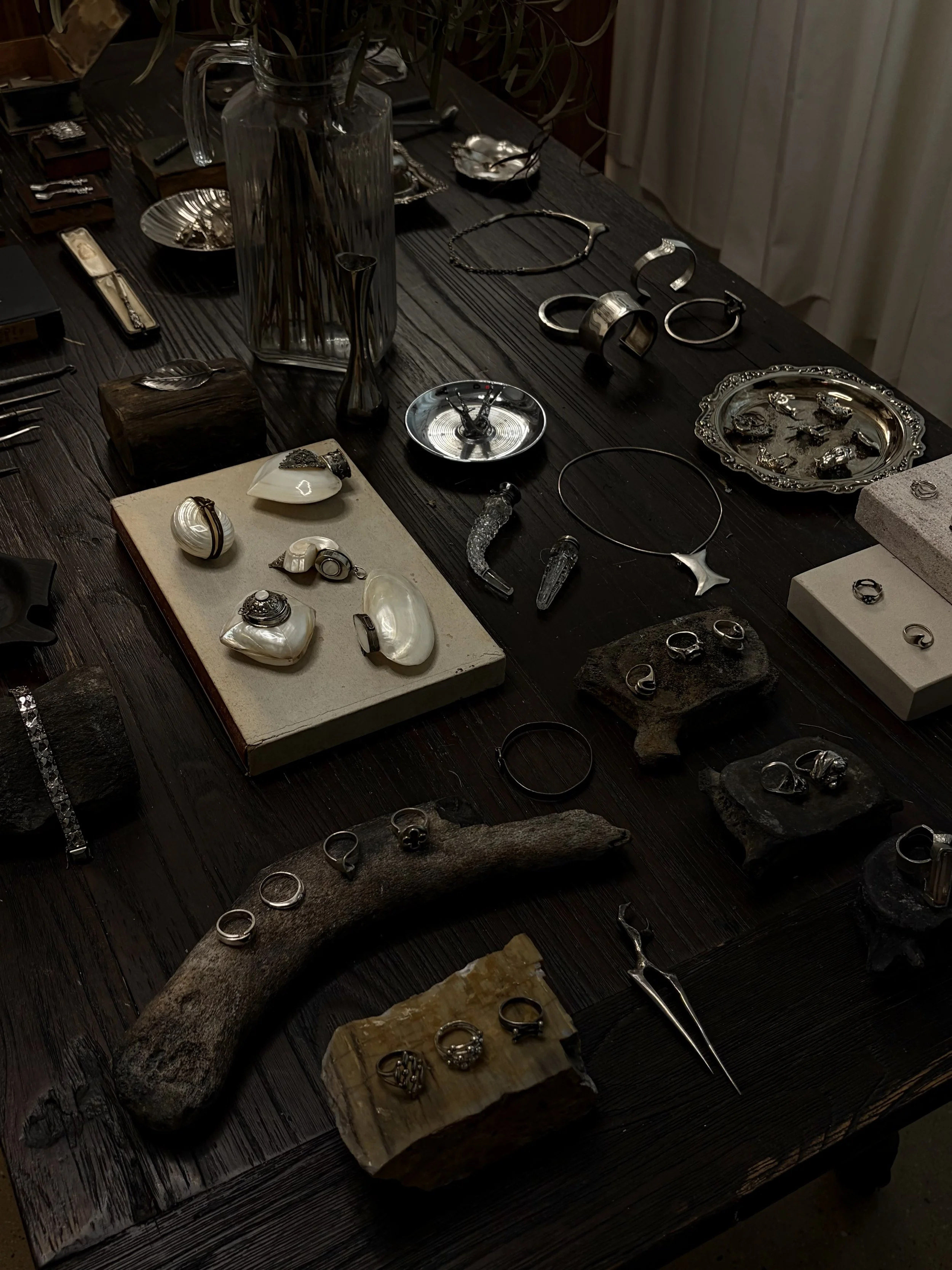Silver jewelry including rings, bracelets, necklaces, and watches displayed on wood and stone surfaces, with a large glass vase and decorative items visible in the background.