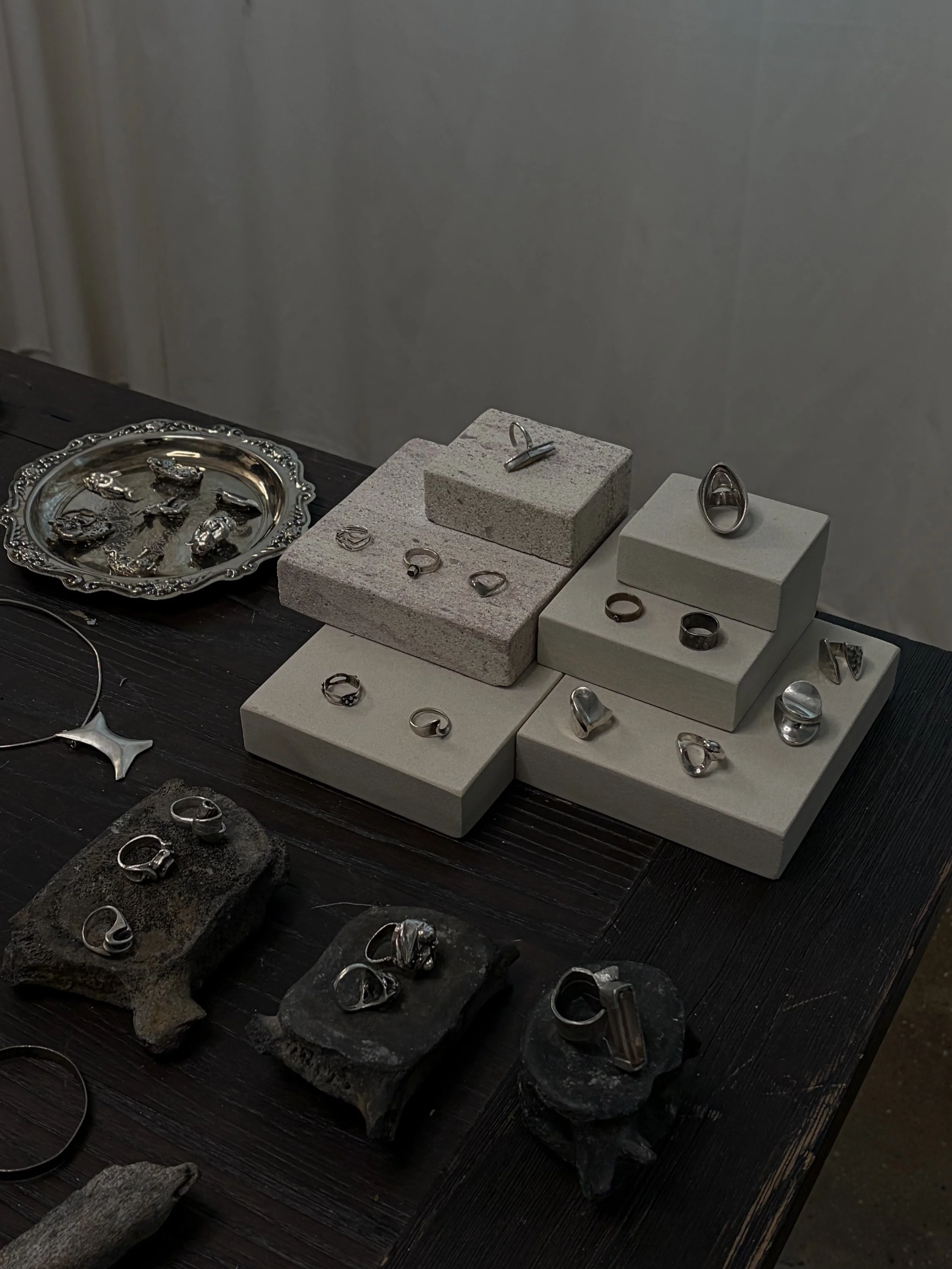 Display of silver jewelry, including rings, earrings, and pendants, arranged on stone slabs and a table, with some jewelry placed on a decorative silver tray.