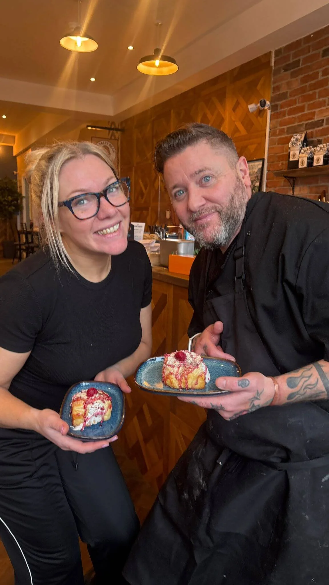 A woman with blond hair, glasses, and a black shirt, and a man with a beard, tattoos, and black clothing, are smiling and holding dessert plates with a slice of cake topped with raspberries, in a cozy restaurant with warm lighting and brick walls.