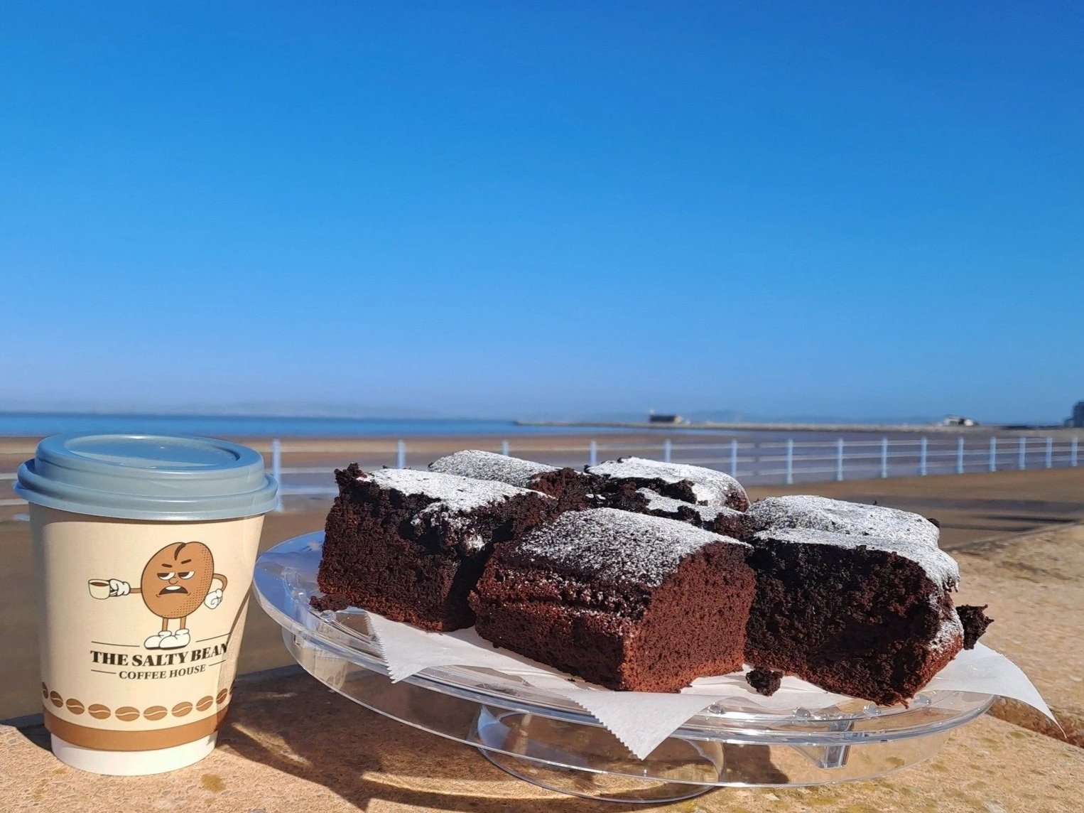 Chocolate cake slices with powdered sugar on a clear plate, a coffee cup with a cartoon cookie character and a blue lid, set outdoors near a beach with a fence, blue sky, and distant boats.