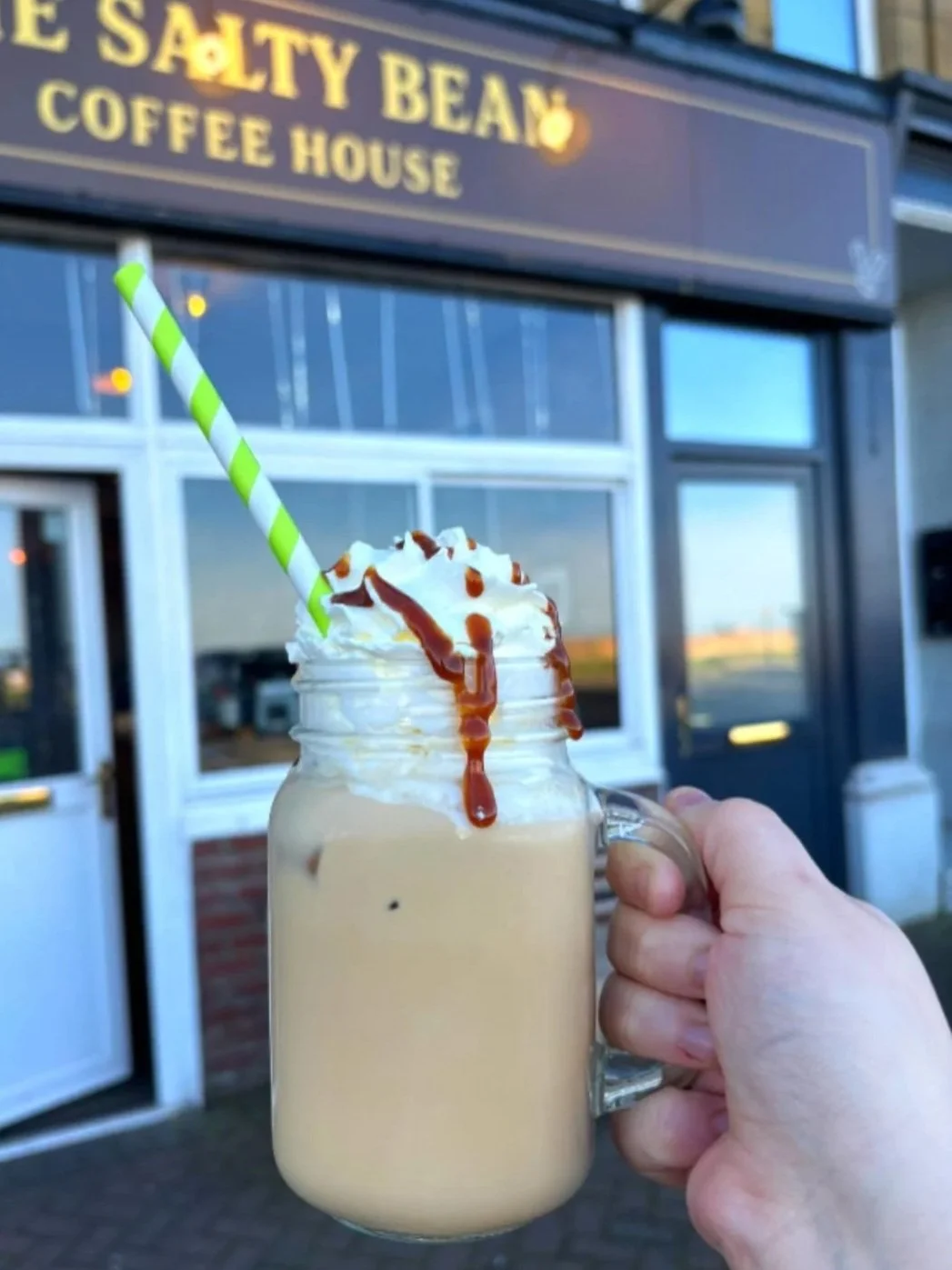 Hand holding a mason jar filled with a creamy milkshake topped with whipped cream, caramel syrup, and a green and white striped straw, in front of the Salty Bean Coffee House storefront.