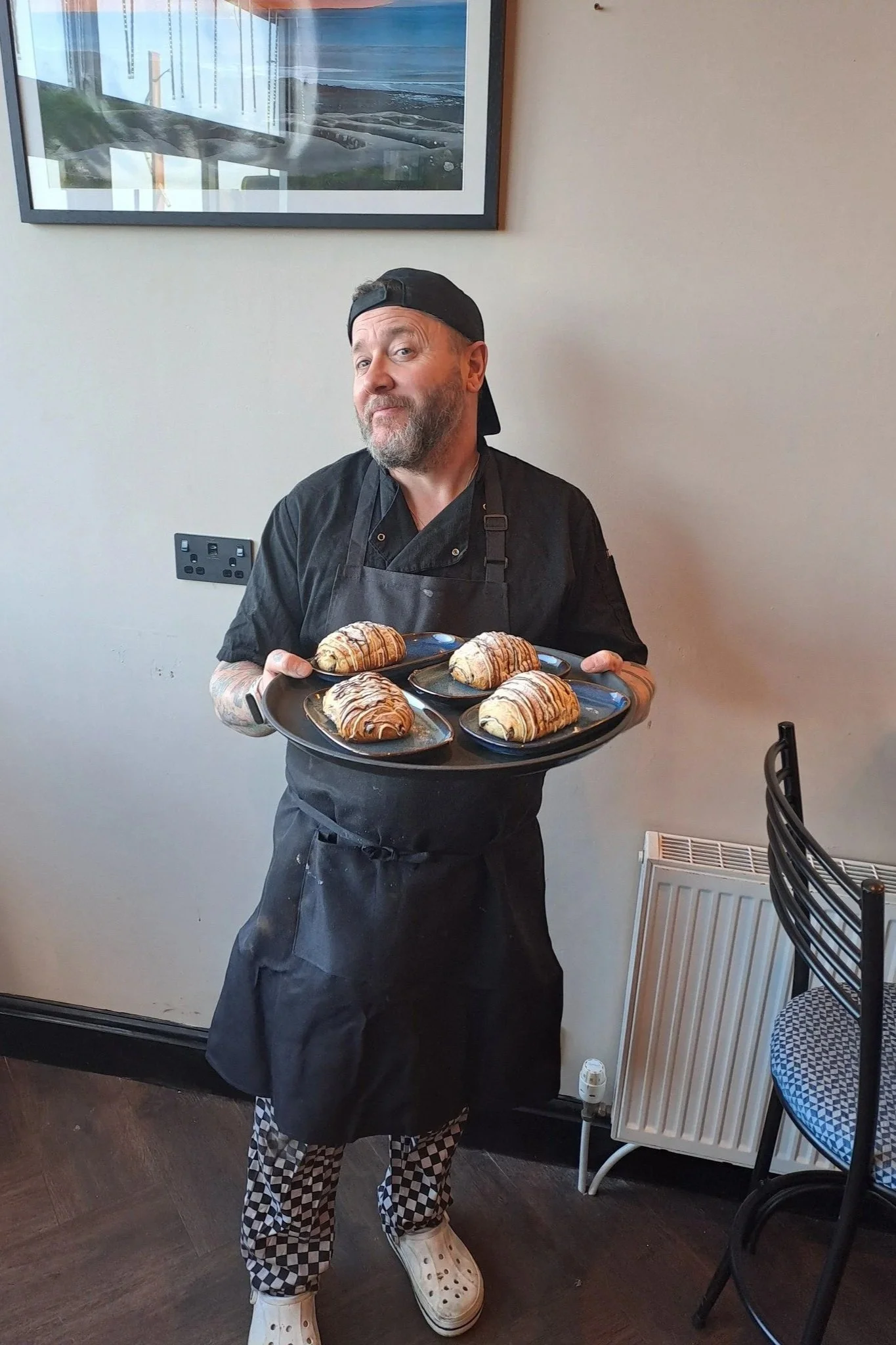 A man wearing a black chef apron, checkered pants, white shoes, a black backward cap, and a wristwatch holding a tray of six croissants.