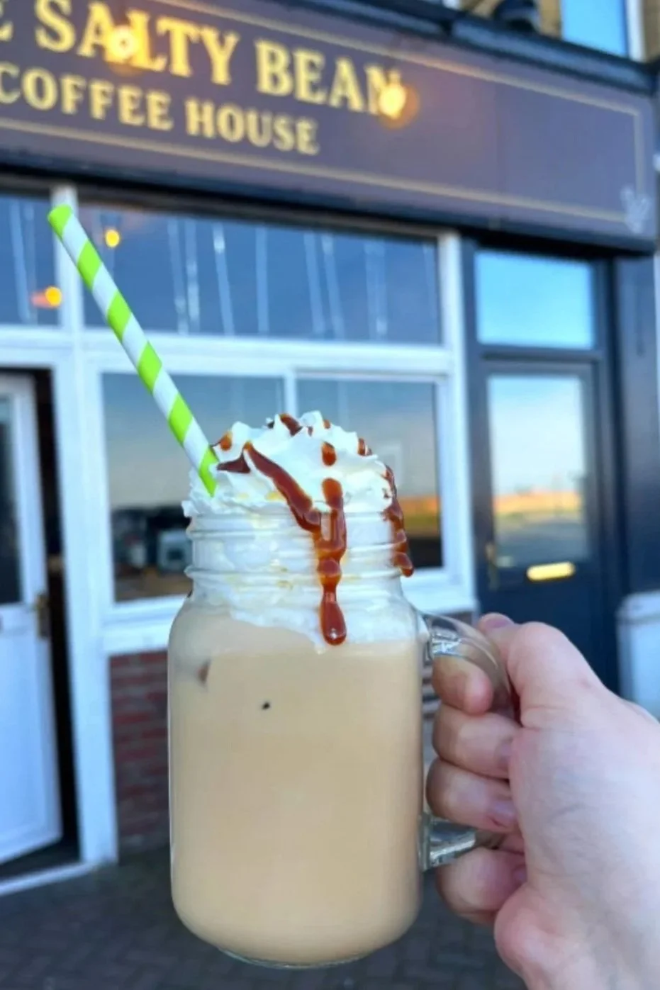 Hand holding a mason jar mug filled with a frappuccino topped with whipped cream, caramel drizzle, and a green and white striped straw outside of a coffee house called 'The Salty Bean Coffee House.'