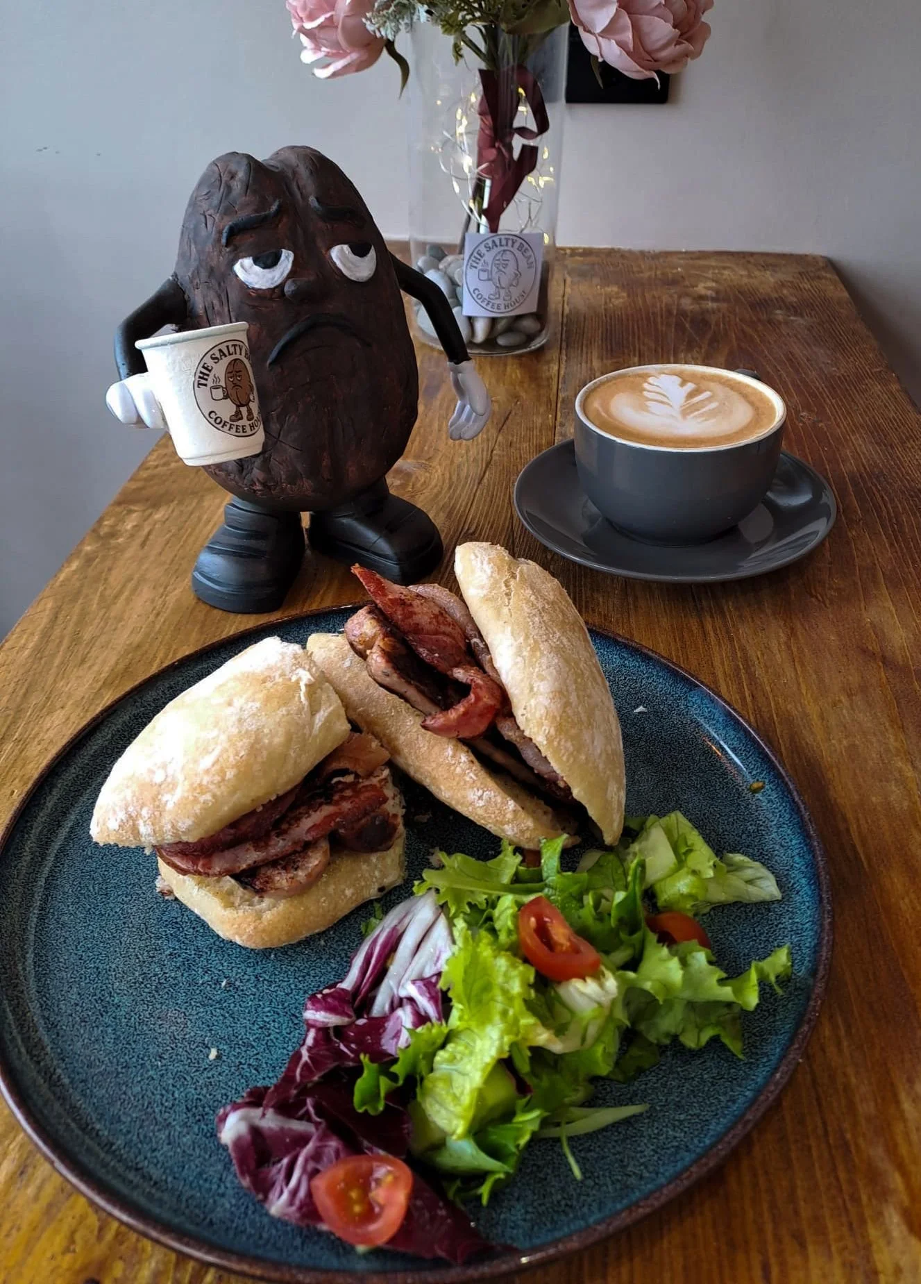 A plate with a sandwich filled with bacon and other ingredients, and a side salad with cherry tomatoes, lettuce, and radicchio. In the background, there is a cup of coffee with latte art, and a coffee cup with a sad face and the text "The Salty Bean Coffee House". There is also a decorative wooden figure holding a coffee cup, and a vase with pink flowers.