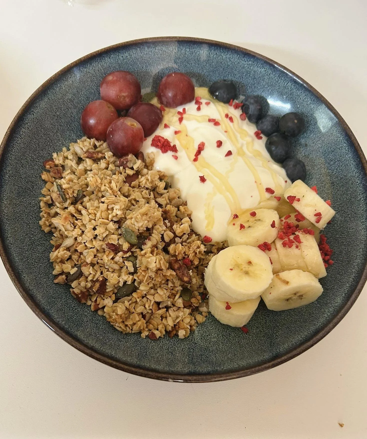A bowl of mixed fruit and granola, including red grapes, blueberries, banana slices, and granola with seeds, topped with honey and red sprinkles.