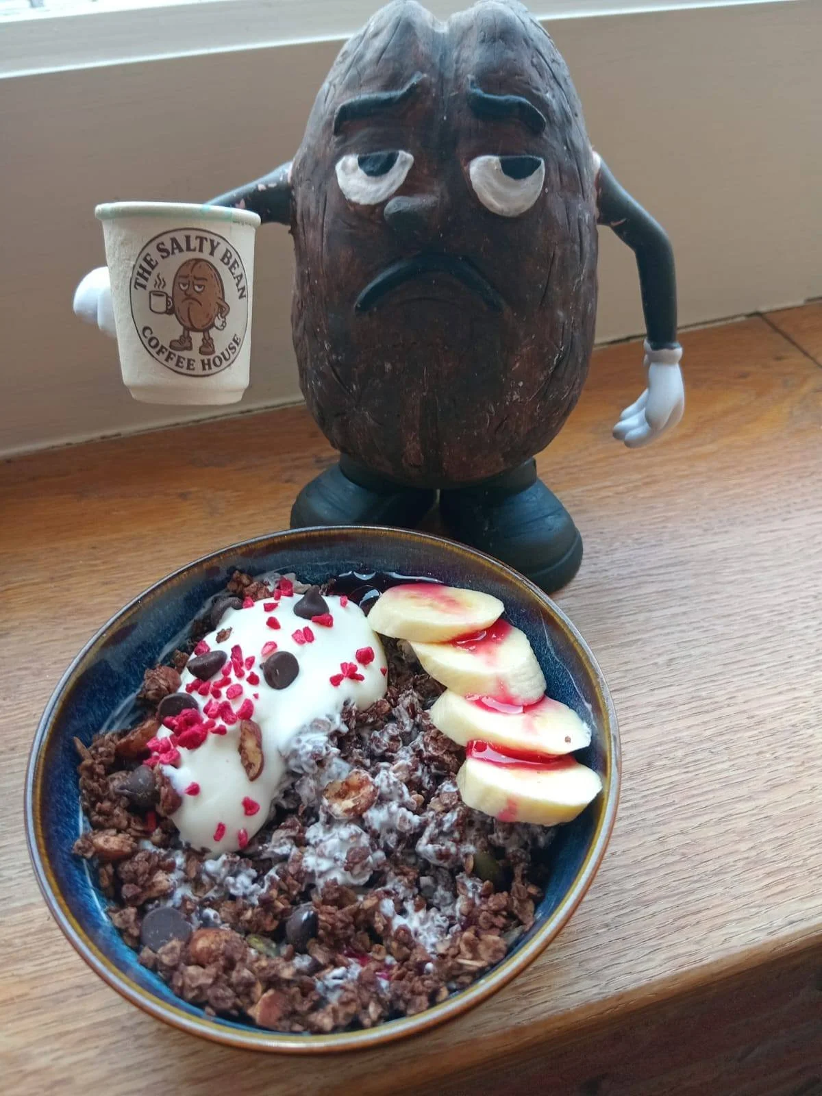 A bowl of yogurt with granola, chocolate chips, red sprinkles, sliced bananas, and syrup. In the background, a wooden mascot figure holding a coffee cup with a sad face and the logo 'The Salty Bean Coffee House'.