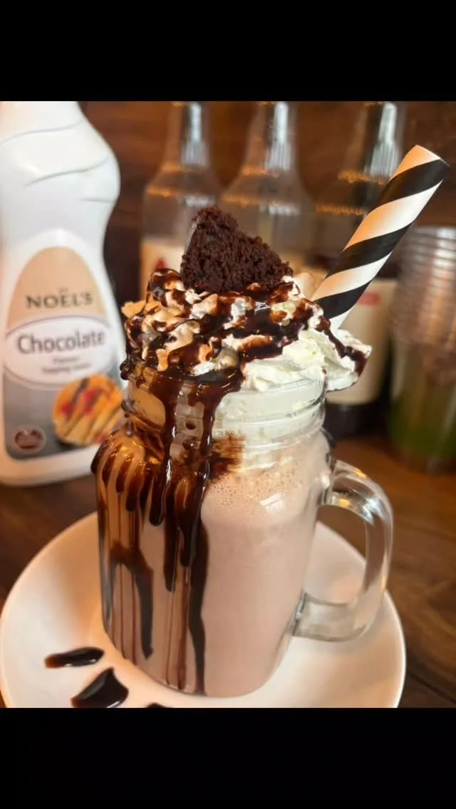 Chocolate milkshake topped with whipped cream, chocolate syrup, and a chocolate cake piece in a glass jar with a black and white striped straw, served on a white plate.