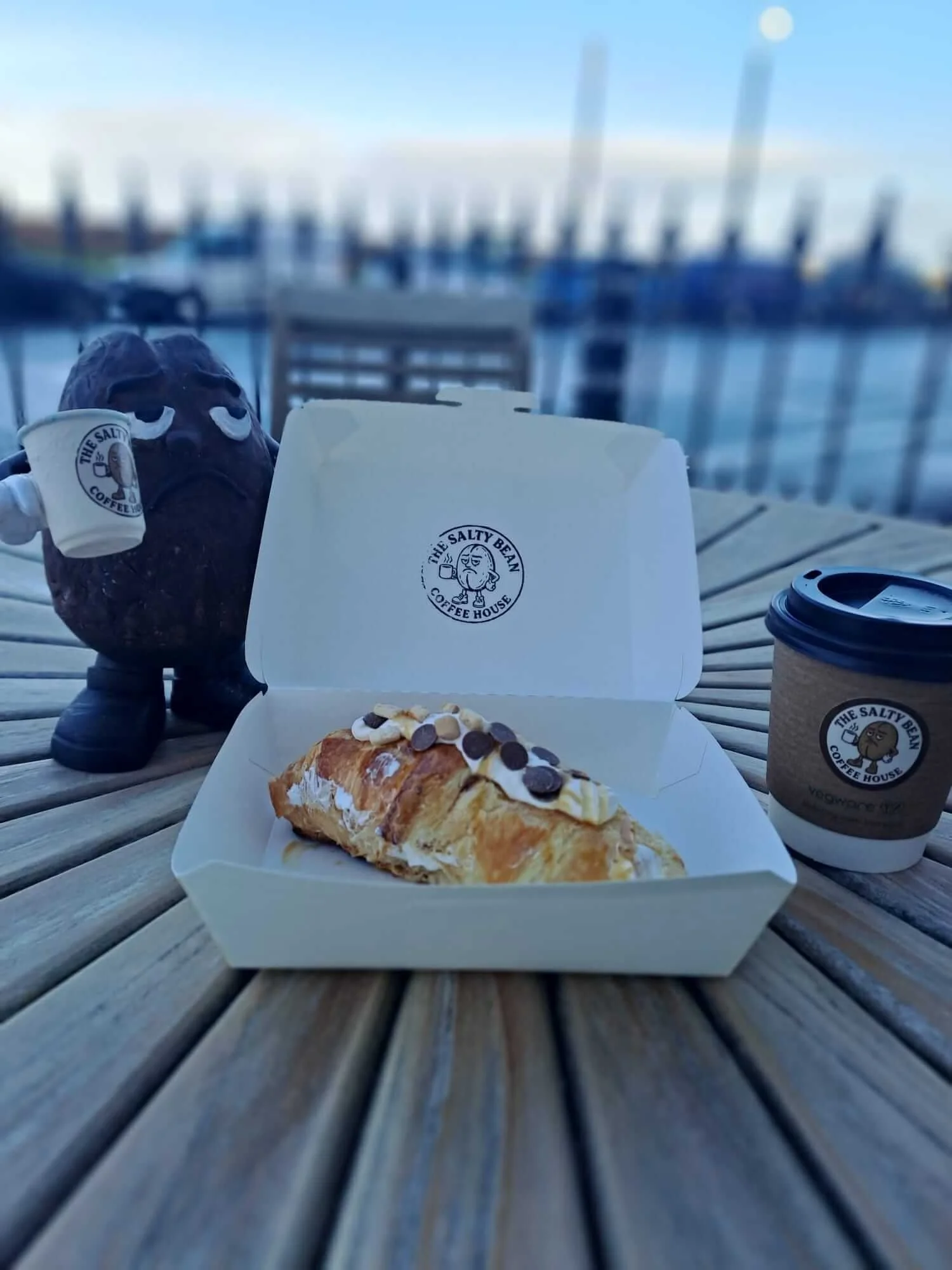 A lunch spread on a wooden table featuring a chocolate croissant with toppings, a coffee cup, and a to-go box with the logo of The Salty Bean Coffee House, with a marina and boats in the background.