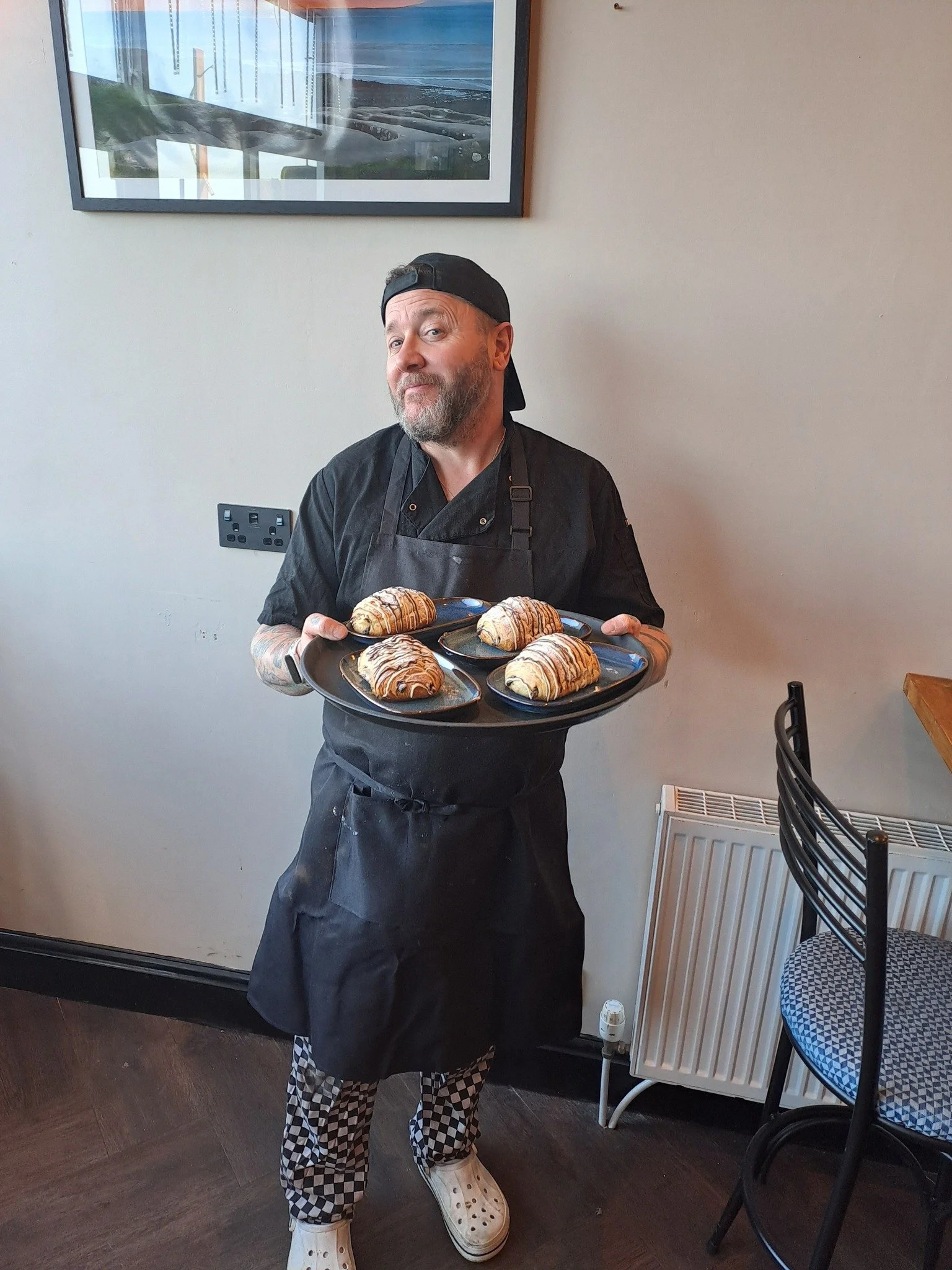 A man with a beard, wearing a black cap backwards, black chef's coat, and checkered pants, is holding a tray with several croissants drizzled with chocolate. He is standing in a room with a light-colored wall, a framed picture, and a radiator, near a
