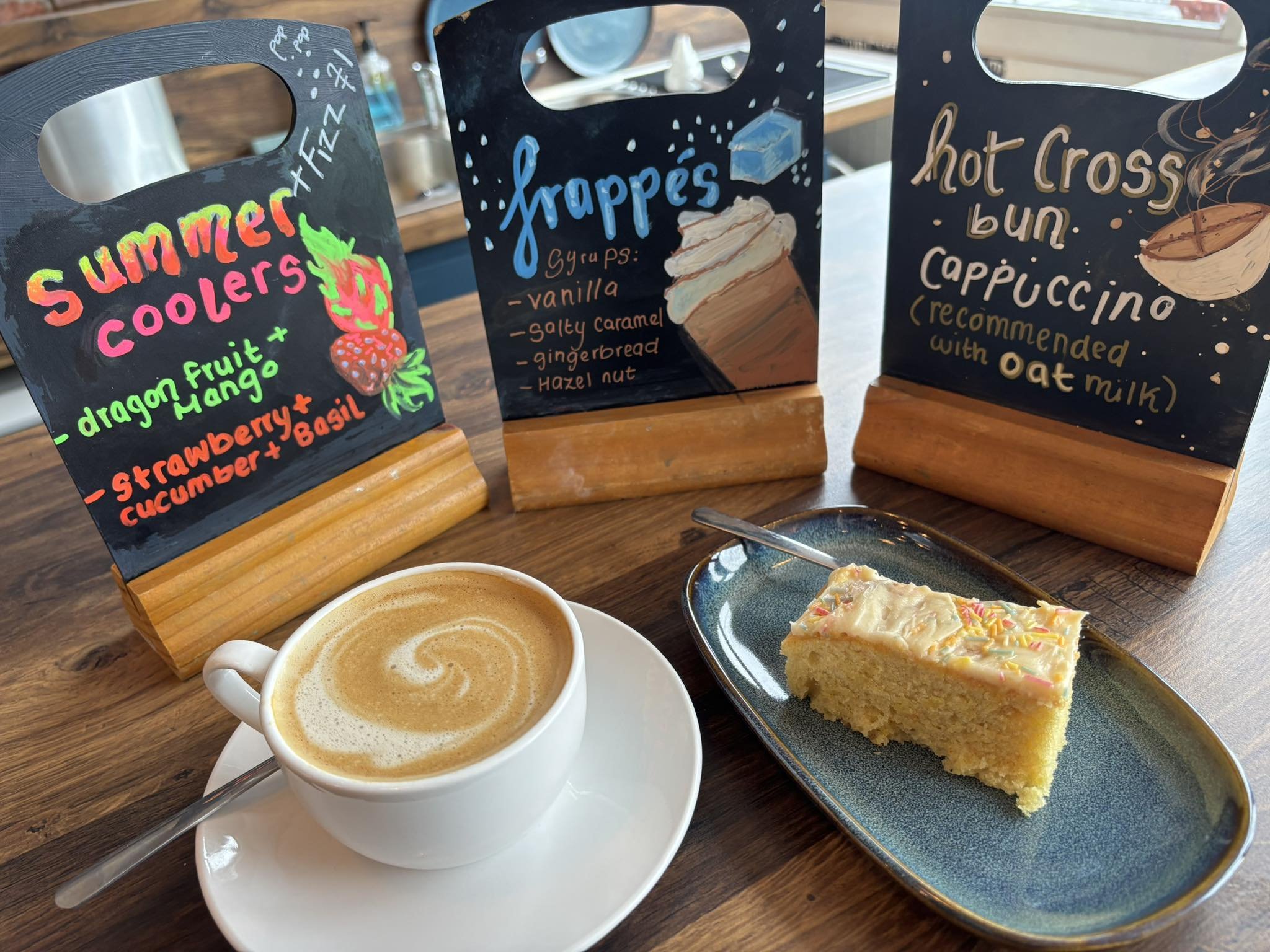 Three chalkboard-style signs with colorful lettering and illustrations sit next to coffee and a slice of cake on a wooden table.
