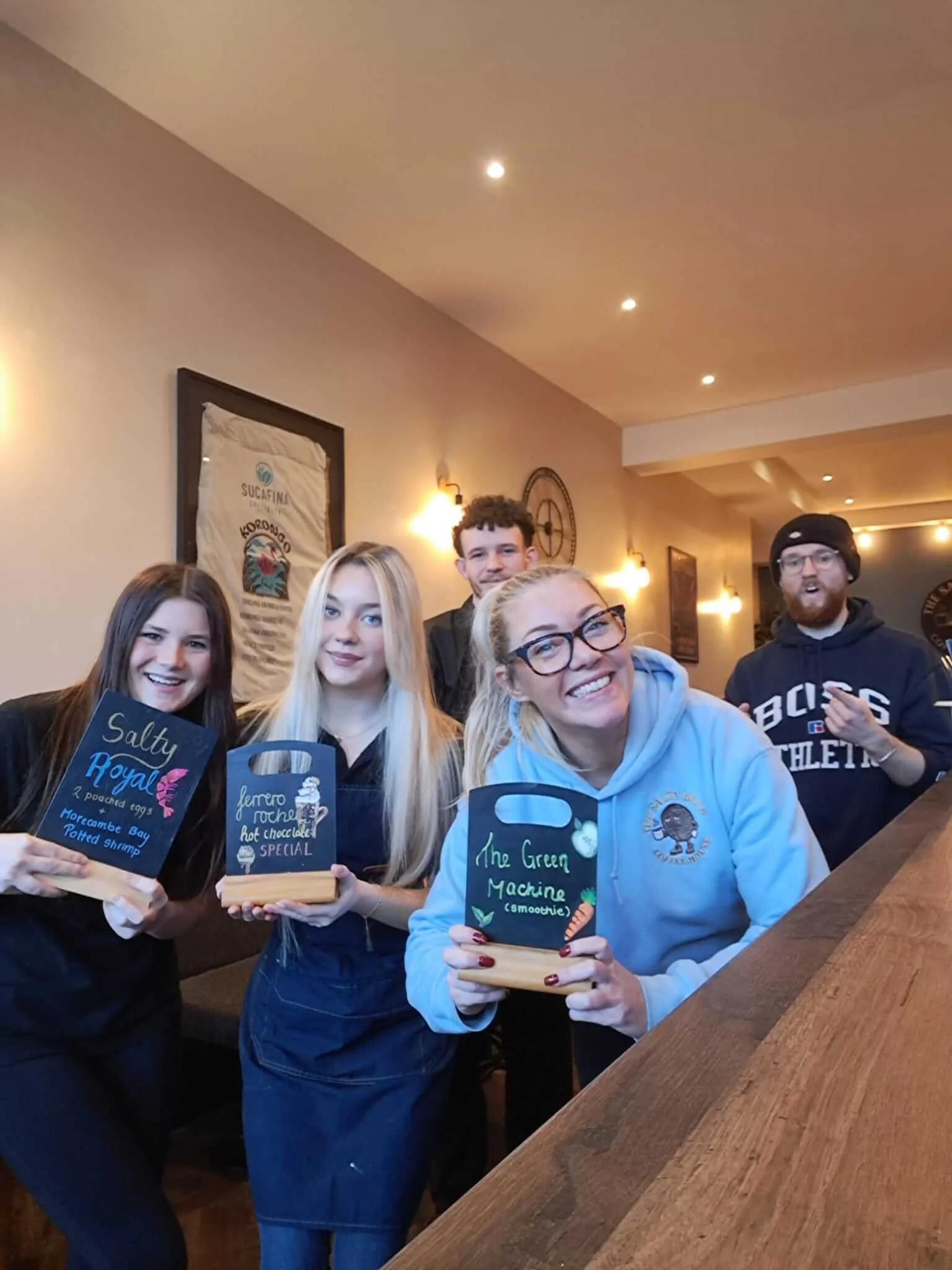 Group of smiling young women and men in a cozy restaurant, holding menu boards and smiling at the camera.