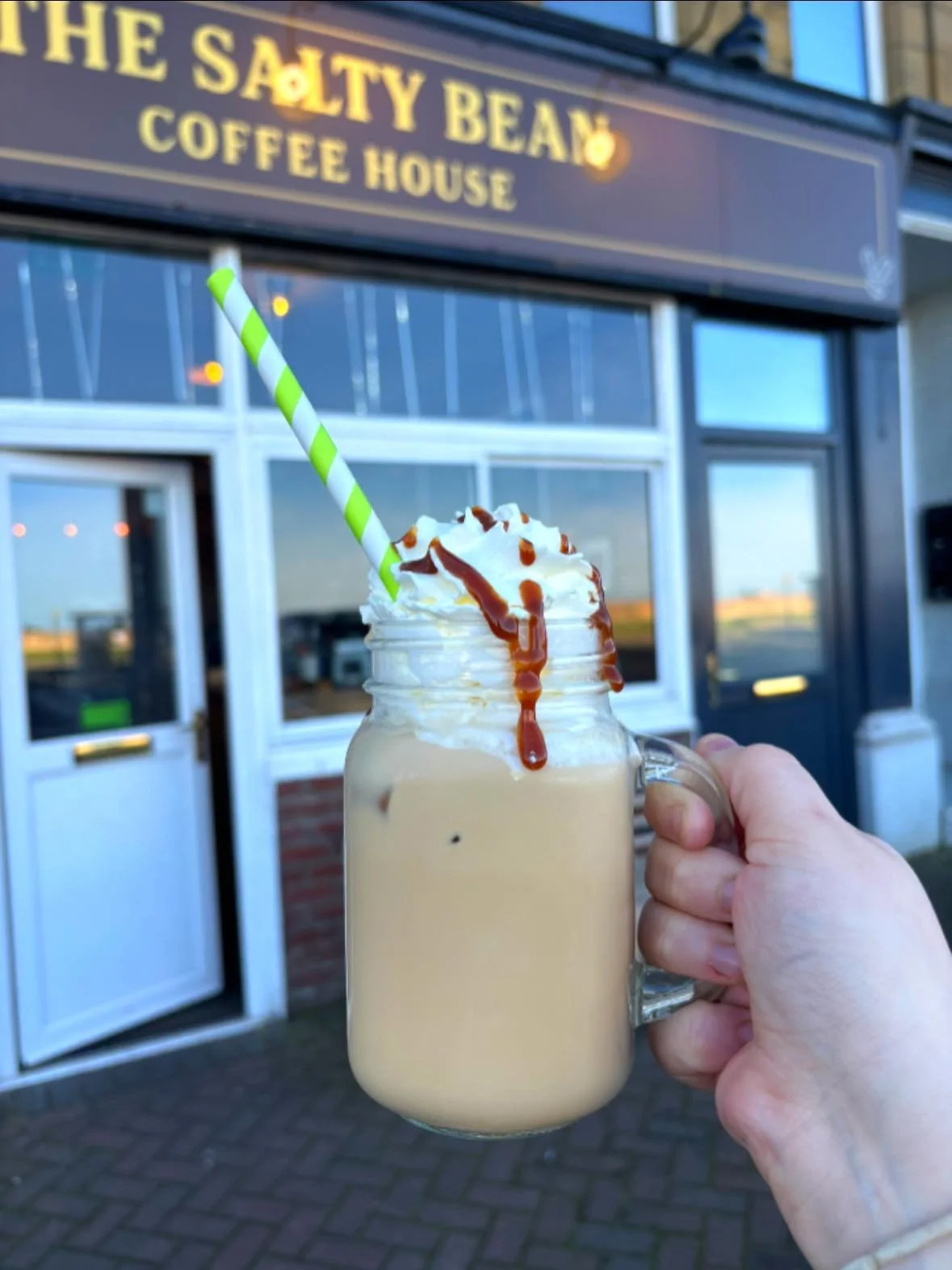 A person holding a glass jar mug of iced coffee topped with whipped cream, caramel, and a green-and-white striped straw, in front of a coffee shop named 'The Salty Bay' in the evening.