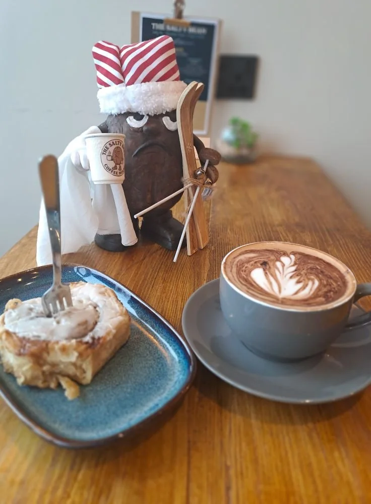 A holiday-themed coffee shop scene featuring a plush coffee mug character wearing a Santa hat, holding a coffee cup, with a latte in a grey cup and a slice of cinnamon roll on a blue plate on a wooden table.