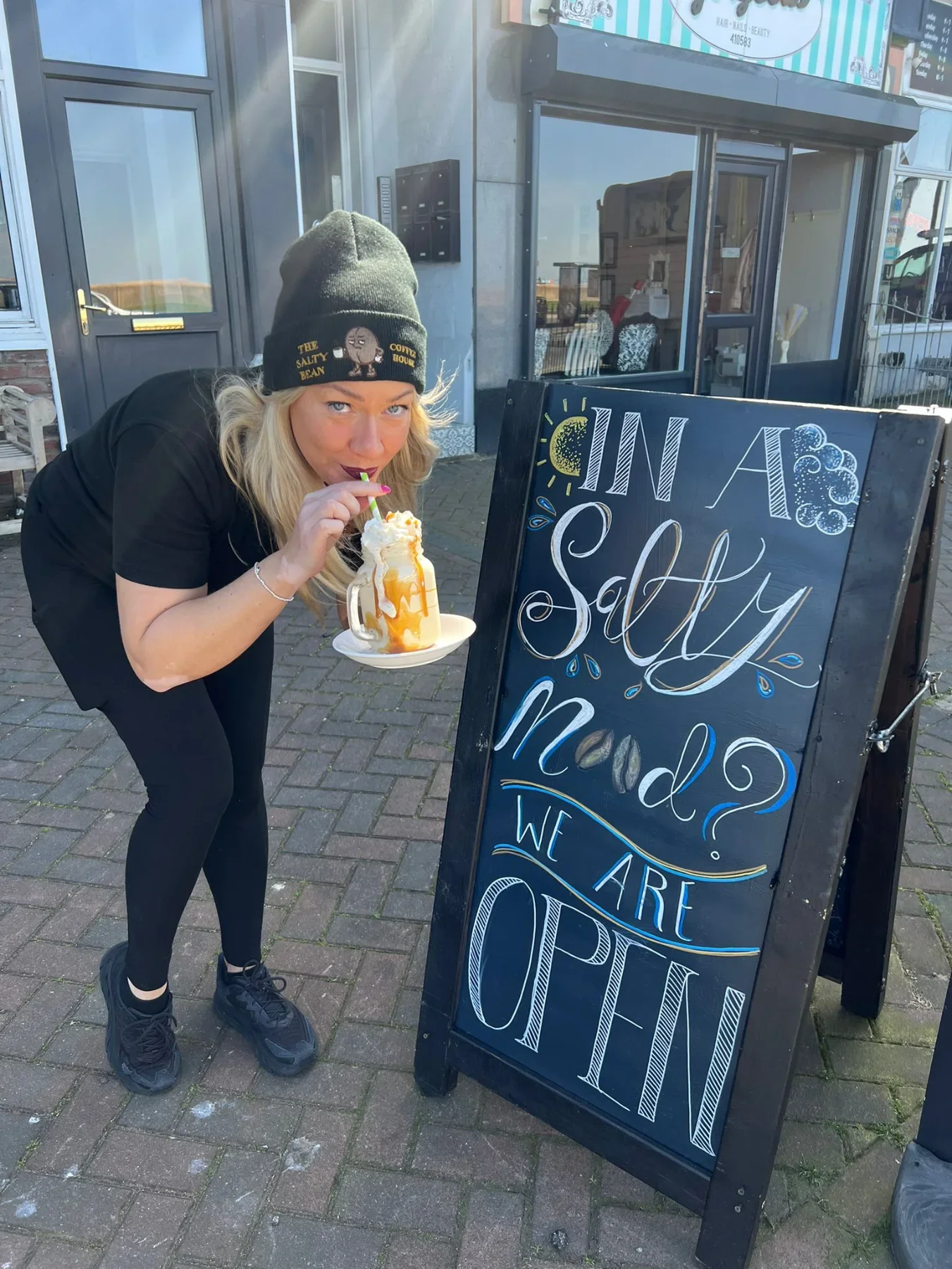 A woman with blonde hair, wearing a black beanie and black athletic clothing, is drinking a large milkshake with whipped cream and caramel sauce outside a shop. She is leaning over a black chalkboard-style sign that reads 'In a salty mood? We are open.' The background shows a storefront and sidewalk.