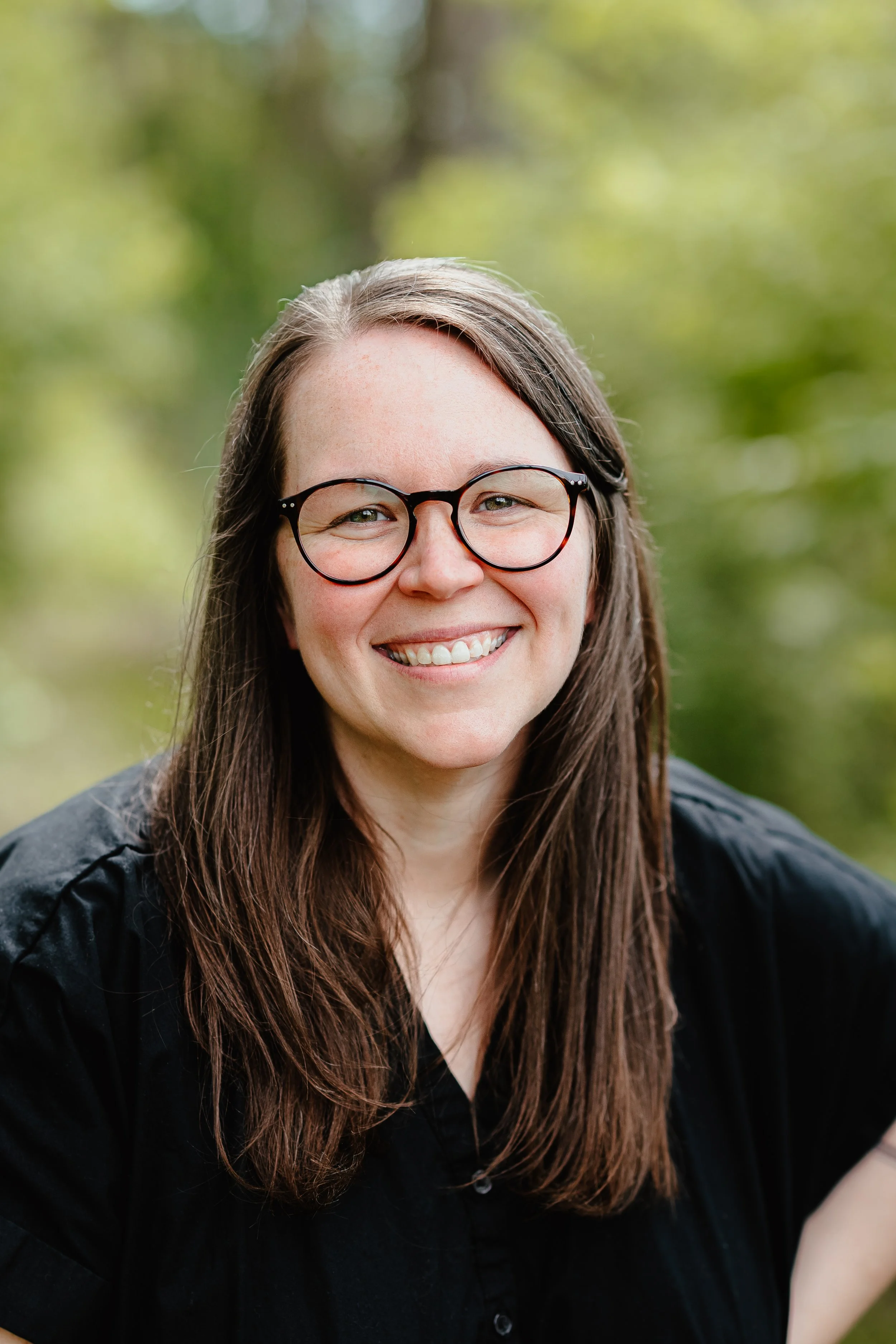 A smiling woman with long brown hair, wearing glasses, outdoors with a blurred green background.