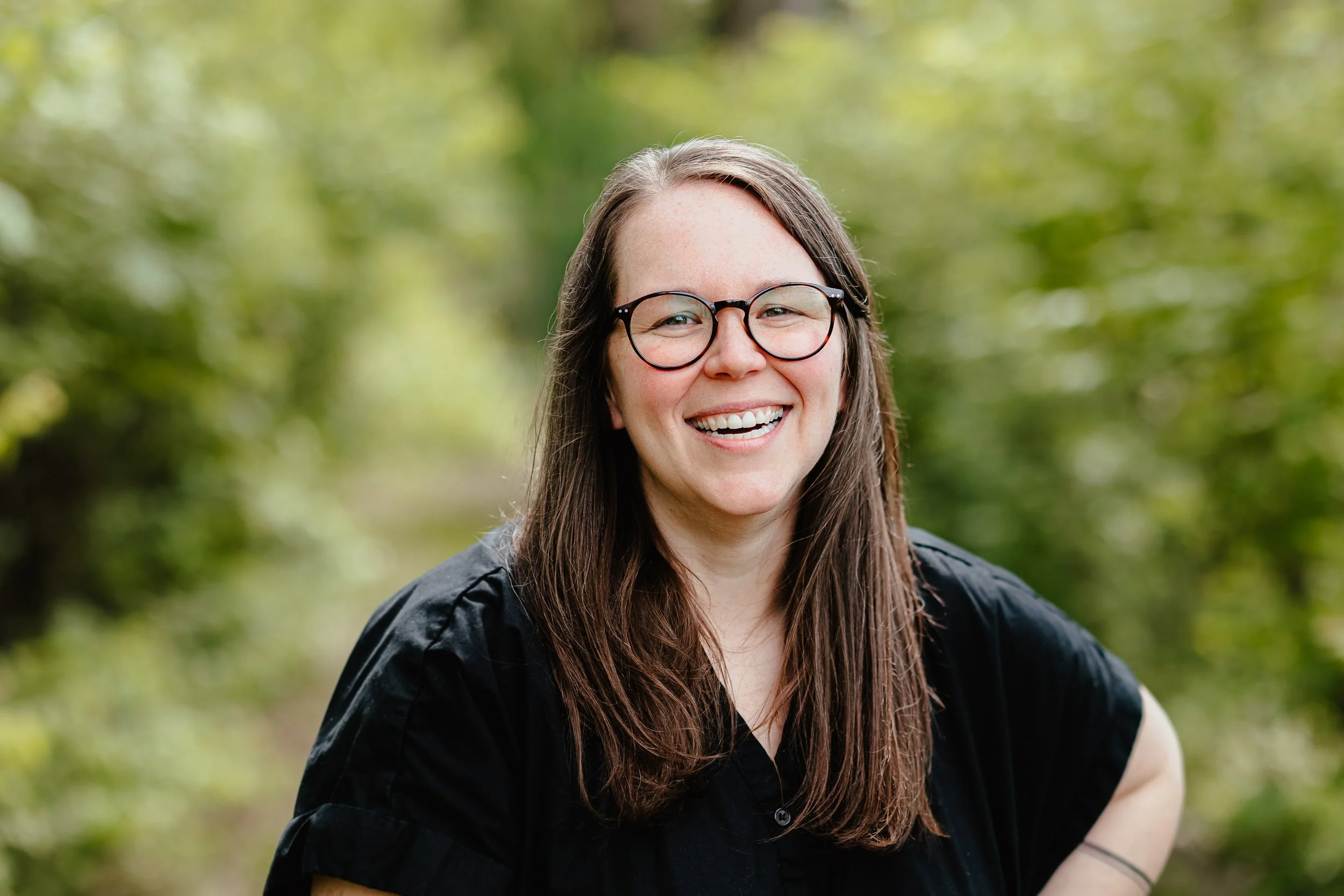 A woman with long brown hair and glasses outdoors, smiling with a green leafy background.