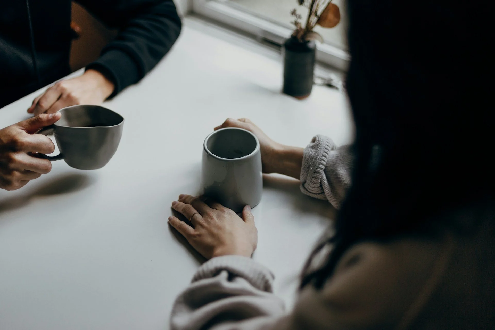 Zwei Personen sind mit Kaffeetassen in der Hand in ein Gespräch vertieft. Sie sitzen an einem weißen Tisch, ein Fenster im Hintergrund, eine Vase mit getrockneten Pflanzen auf dem Fensterbrett.