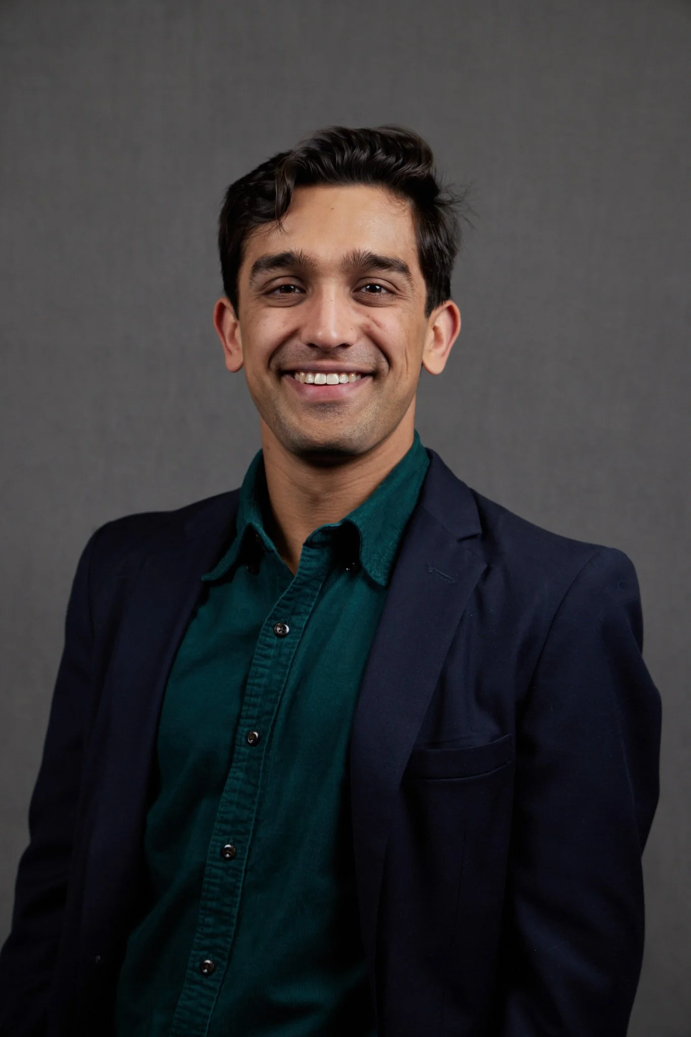 Portrait of a smiling man with dark hair, wearing a green shirt and a dark blazer, standing against a plain grey background.