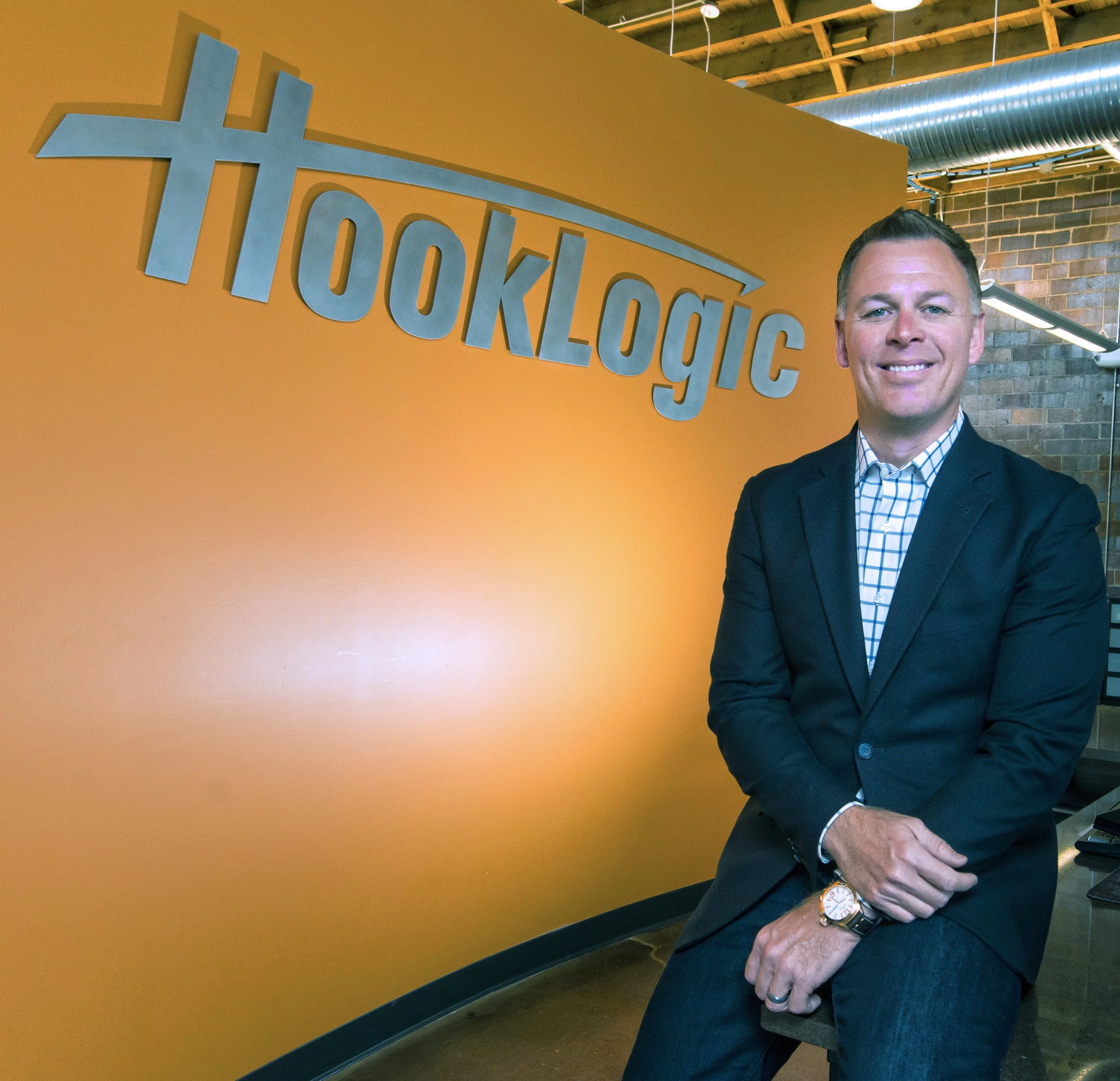 A man in a suit and checkered shirt standing in front of an orange wall with the hashtag #ToughLydic in large blue letters. He is smiling and has short dark hair. The ceiling shows exposed wooden beams and metal ducts.