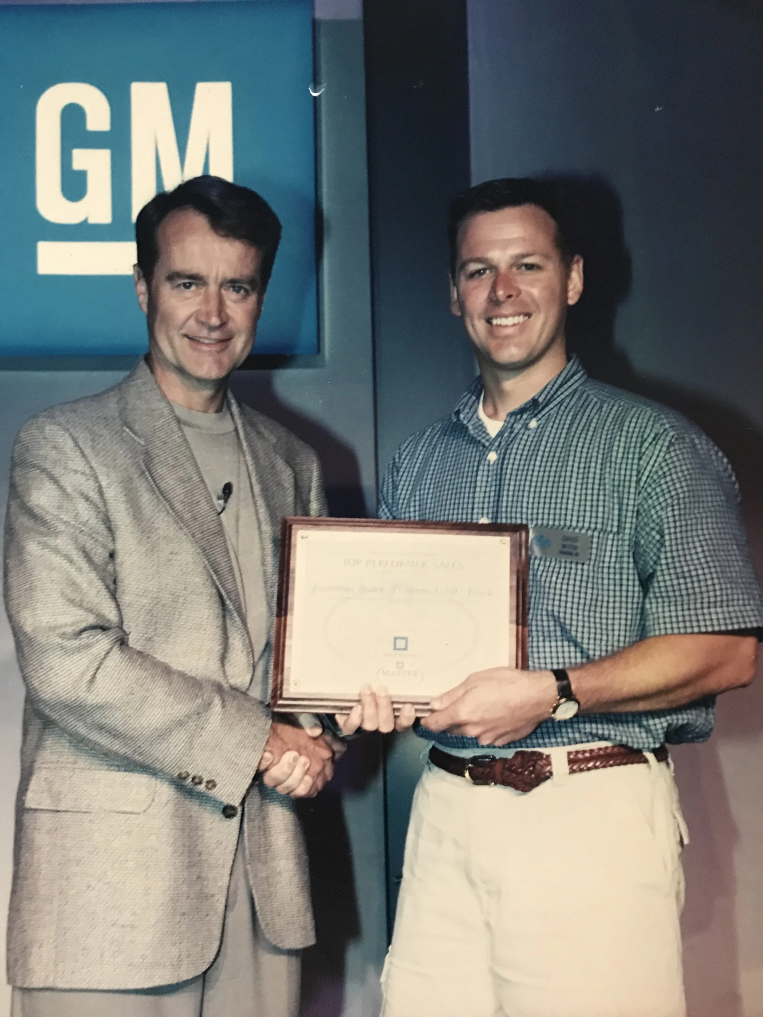 Two men shaking hands, one holding a framed certificate, in front of a GM sign.