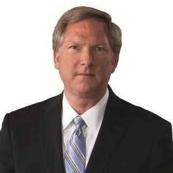 A man with light brown hair wearing a black suit, white shirt, and blue striped tie, posing against a white background.