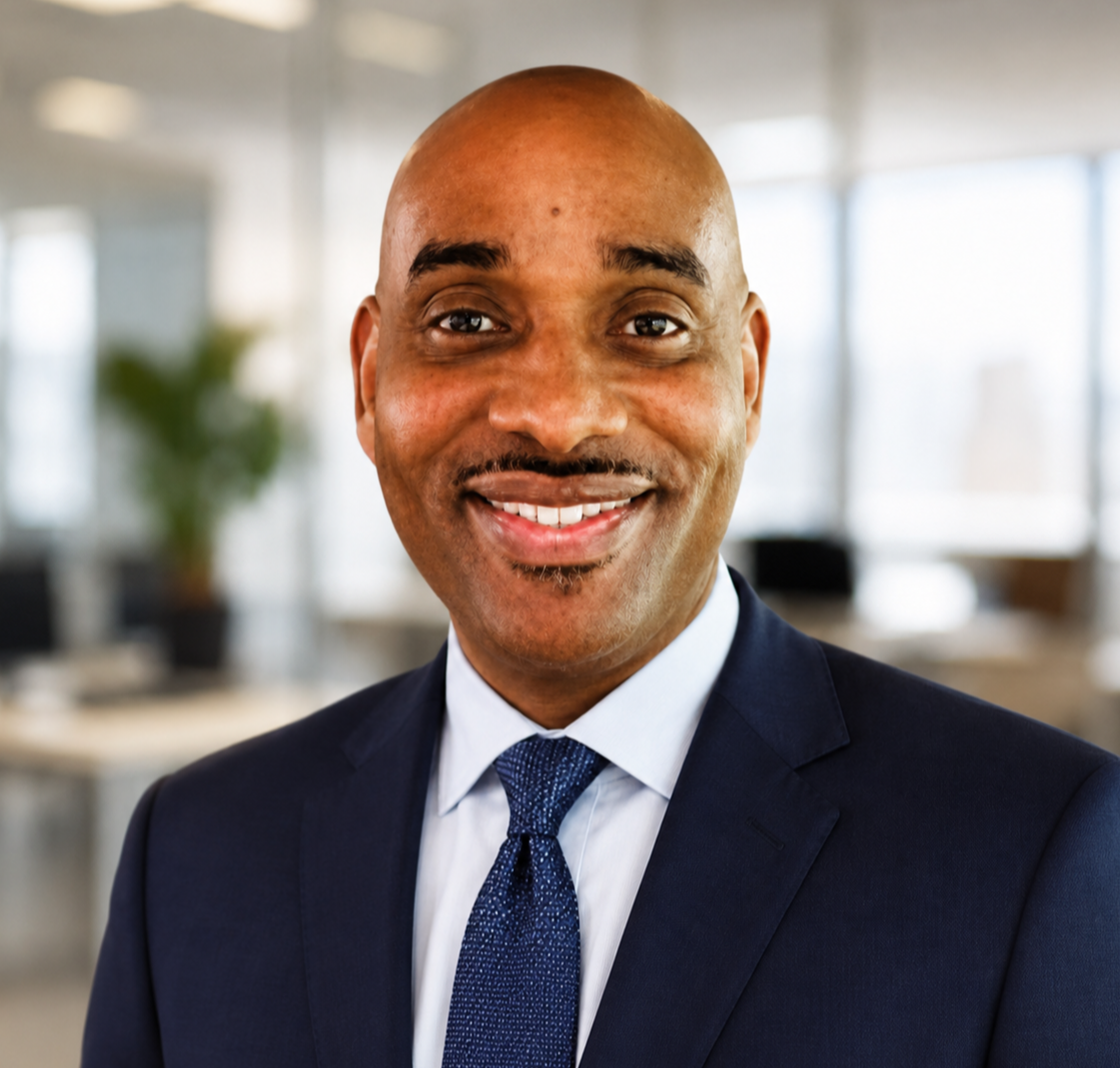 A smiling African American man in a navy suit, white shirt, and blue tie, in an office setting with blurred background of windows and plants.