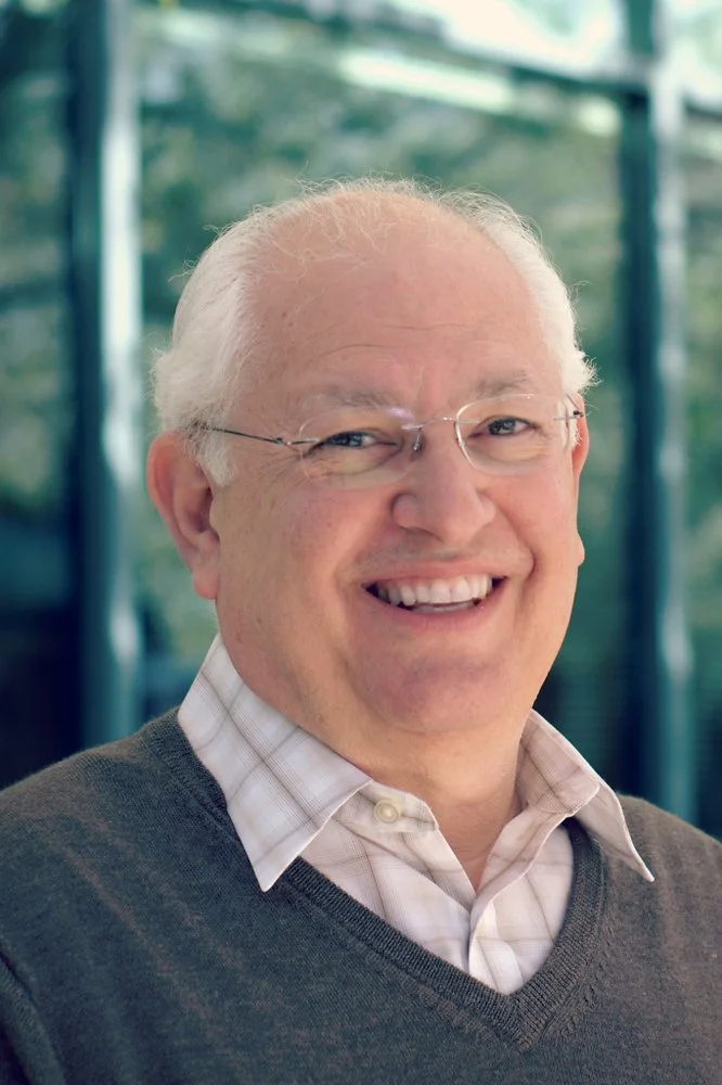Portrait of a smiling man with glasses, white hair, wearing a white collared shirt and a gray sweater, standing in front of a glass building with trees reflected in the background.