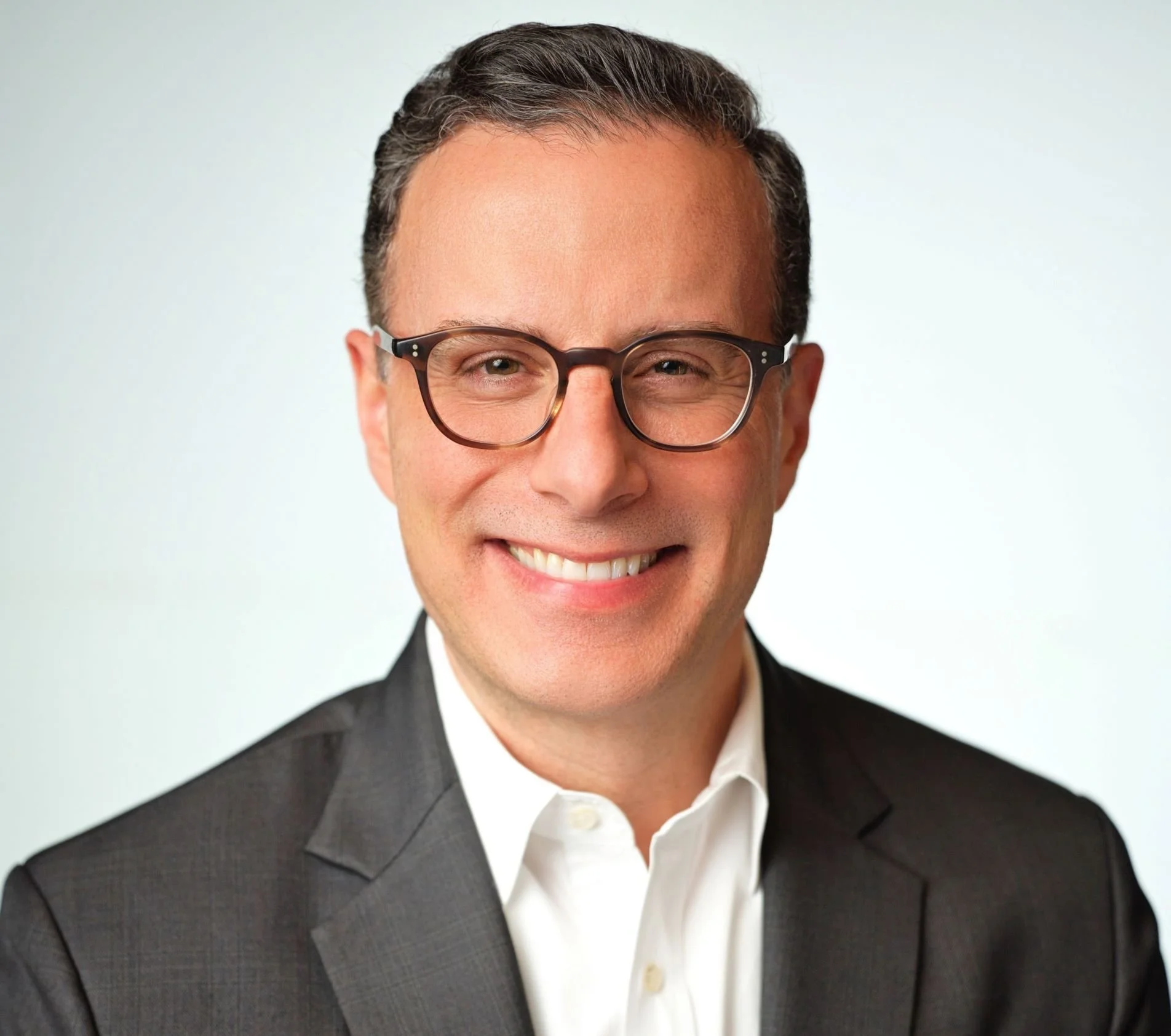 Close-up of a man with dark hair and glasses, wearing a suit and smiling against a plain light background.