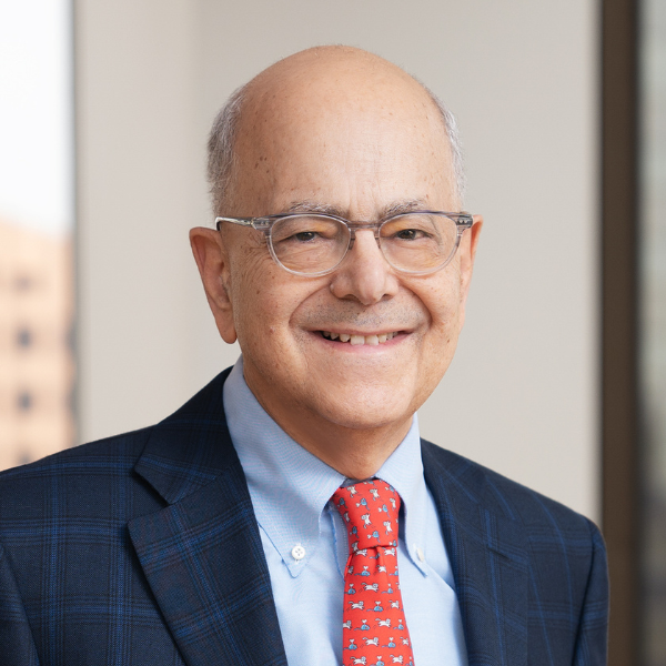 A middle-aged man with glasses, dressed in a checkered navy blazer, light blue shirt, and red patterned tie, smiling in front of a window with a city view.