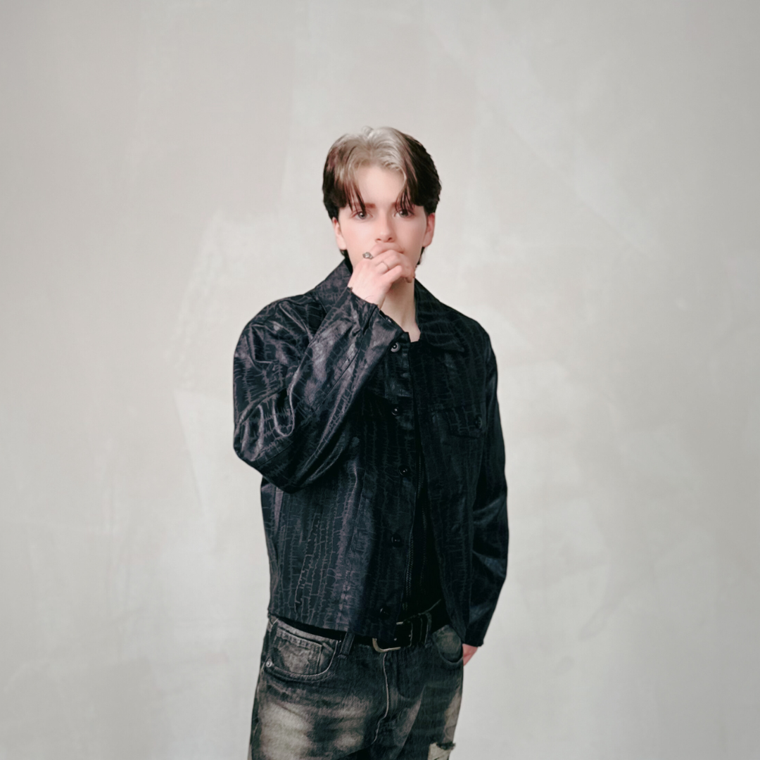 Young man with dark hair wearing a black patterned jacket and jeans, posing against a neutral background.
