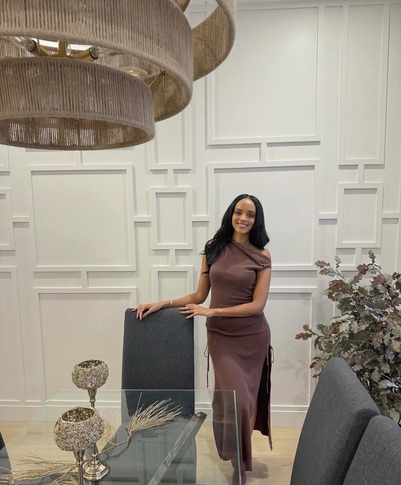 A woman with long black hair in a brown dress standing next to a glass dining table in a modern room with white paneled walls and decorative plants, smiling at the camera.