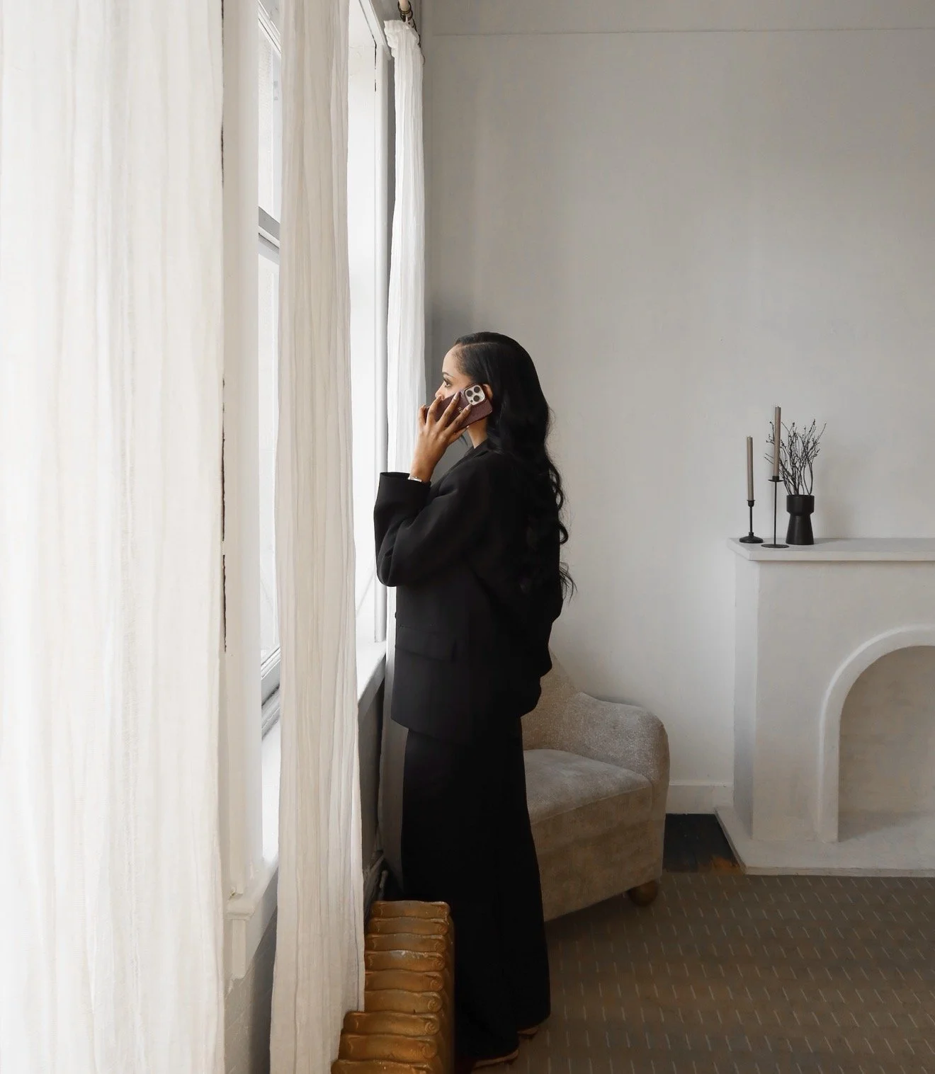 A woman with long dark hair, dressed in black, is talking on a cellphone near a window with white curtains in a minimalistic, modern room.