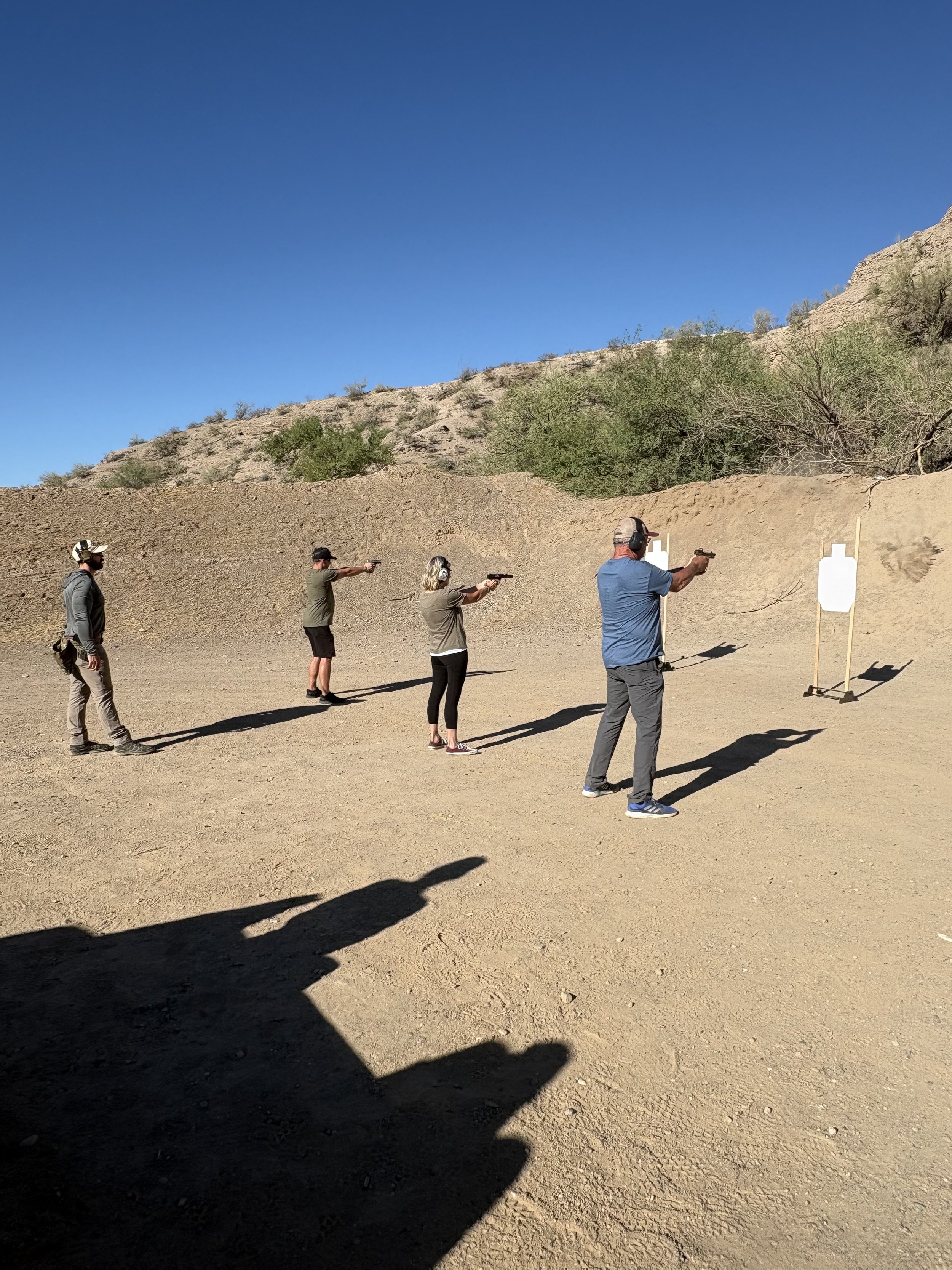 Four people at an outdoor shooting range, aiming handguns at targets, with desert landscape and blue sky in the background.