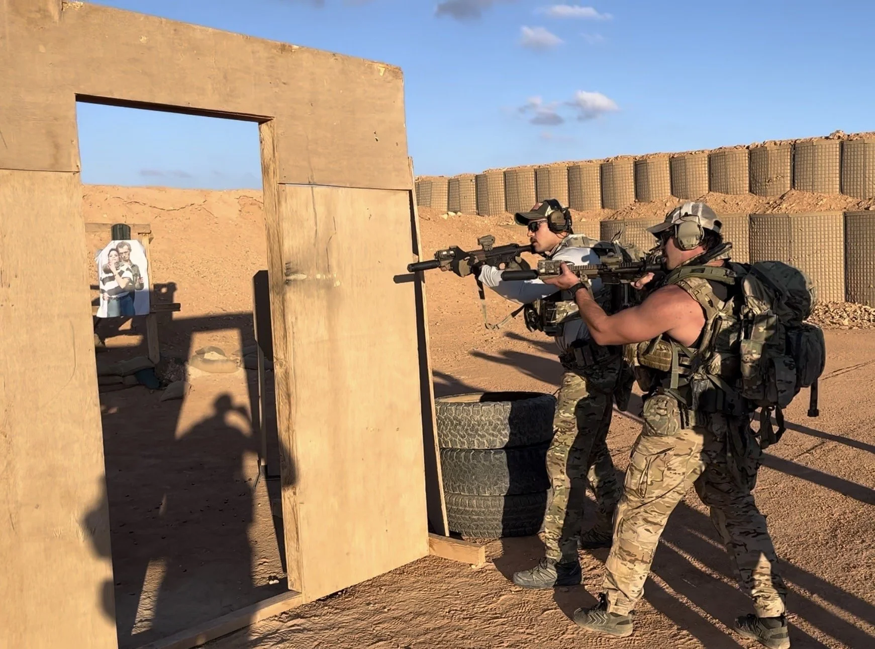 Two men in camouflage military uniforms shooting armed rifles at a shooting range. They are wearing protective ear gear and backpacks, aiming through a cardboard barrier. There is a target with a photo of two women on it.