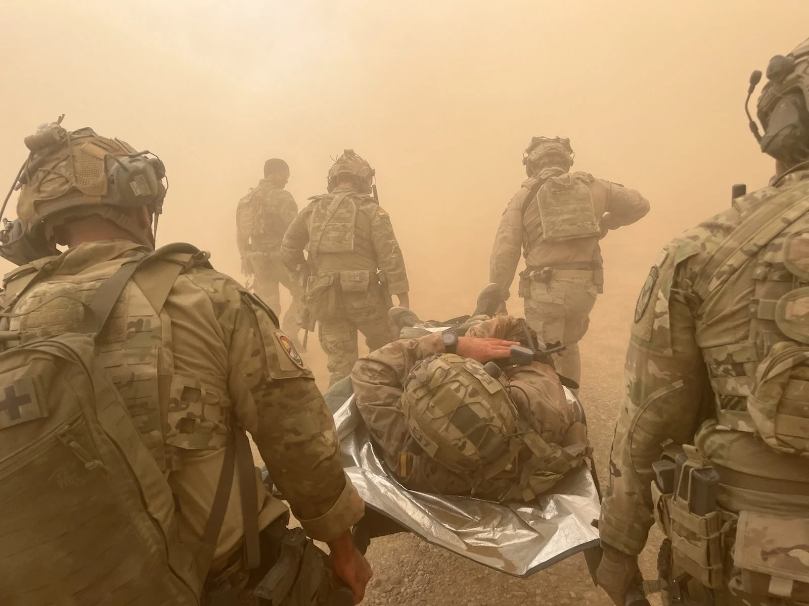 Military personnel rescuing an injured soldier from a dusty environment, with some soldiers attending to him on a stretcher.