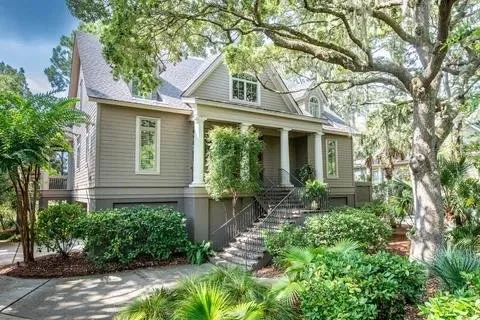 A gray house with a front porch, surrounded by lush green trees and shrubs, with concrete stairs leading to the entrance in a landscaped yard.