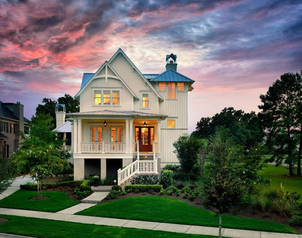 A large two-story white house with a wraparound porch, surrounded by green lawns and trees, under a colorful sunset sky.