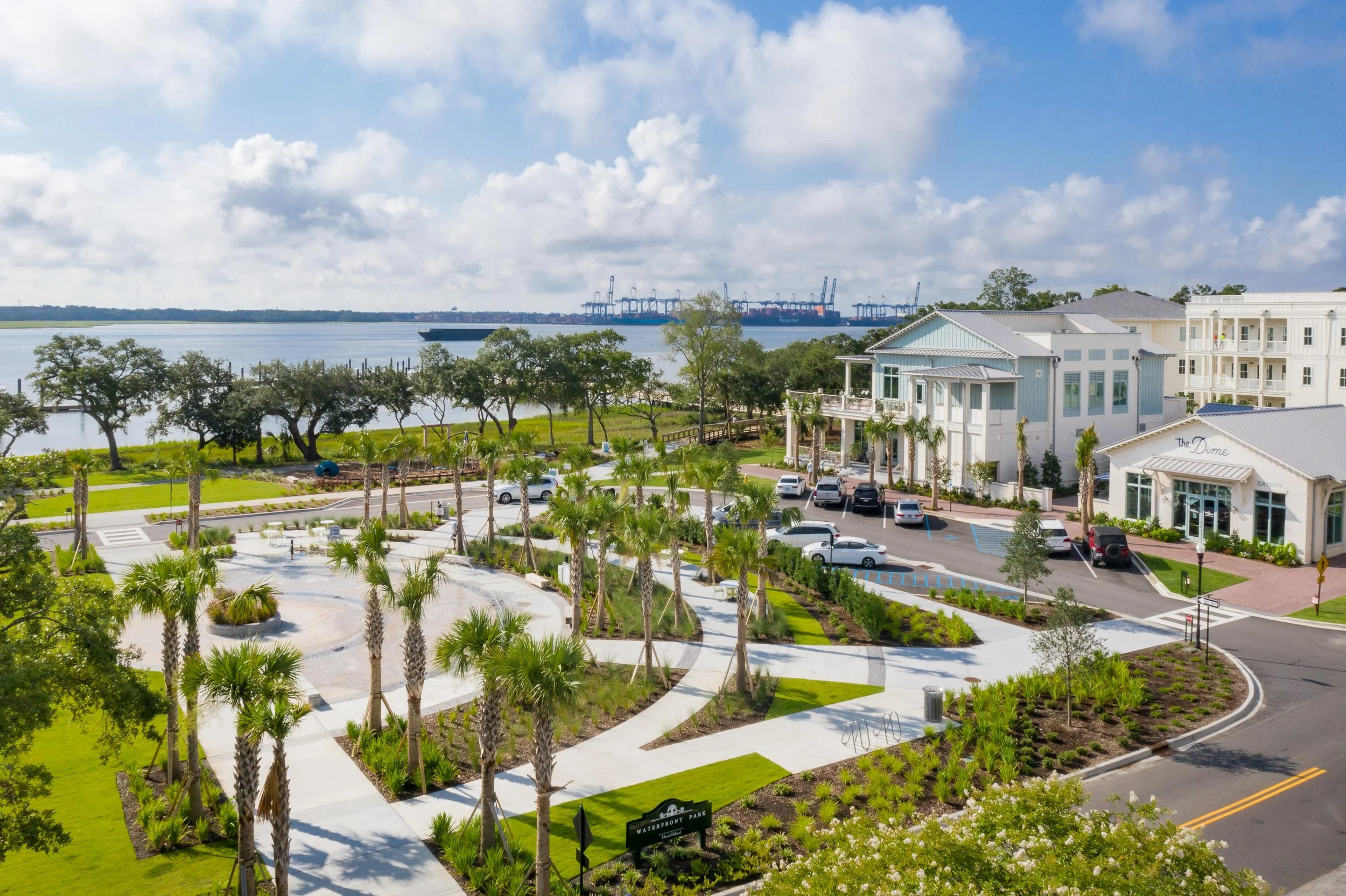 View of a waterfront park with palm trees, paved walkways, and parked cars in the foreground. Buildings are on the right, and a large body of water with a cargo ship and industrial cranes in the distance are visible under a partly cloudy sky.