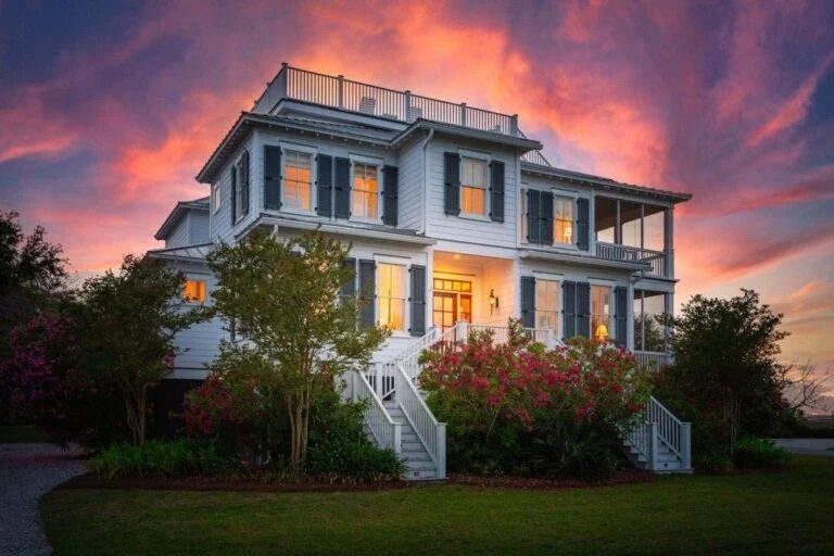 A large white house with multiple stories, black shutters, and a front porch, surrounded by lush bushes and trees, during a colorful sunset.